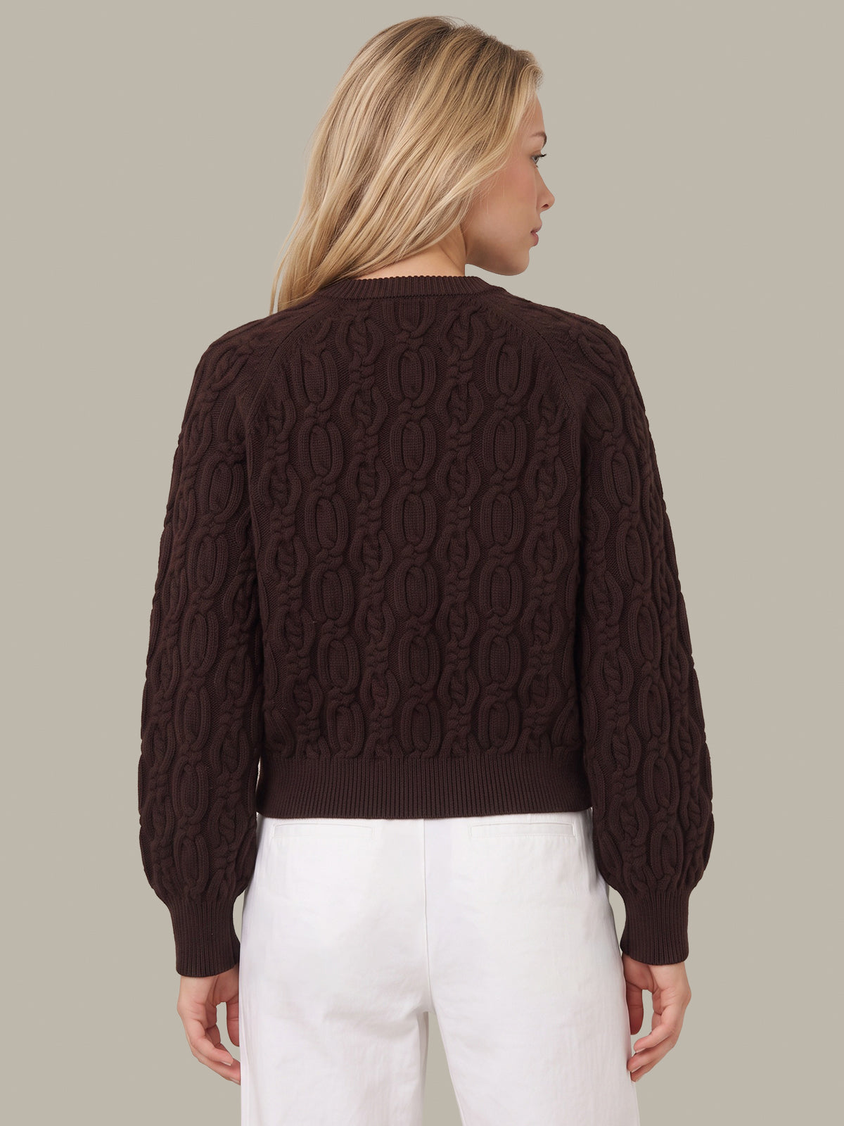A woman shown from the back wearing a dark brown cable-knit sweater and white pants, set against a solid light gray background.