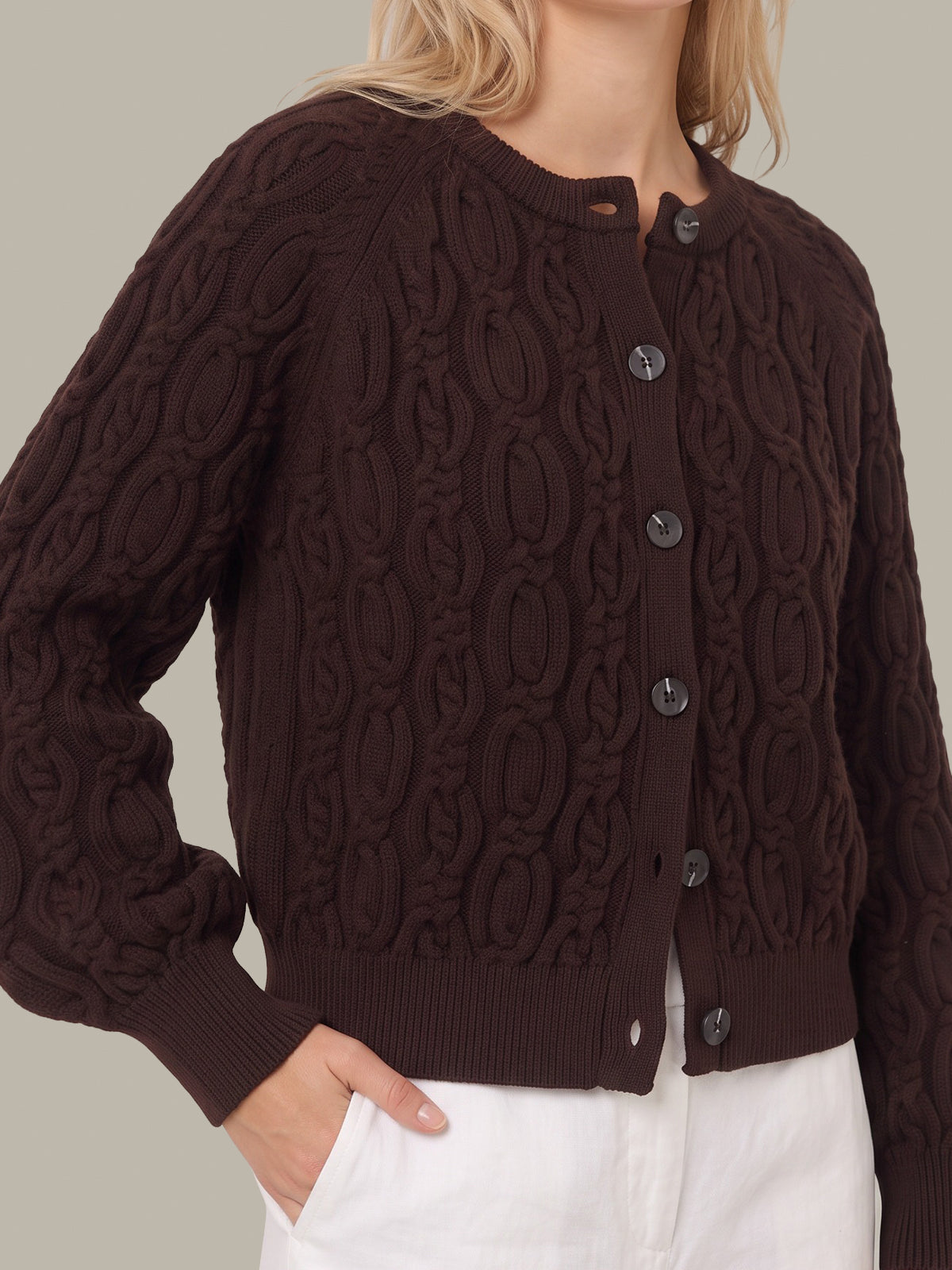 A woman wearing a dark brown cable-knit cardigan sweater with dark buttons, paired with white pants, shown against a solid light gray background.