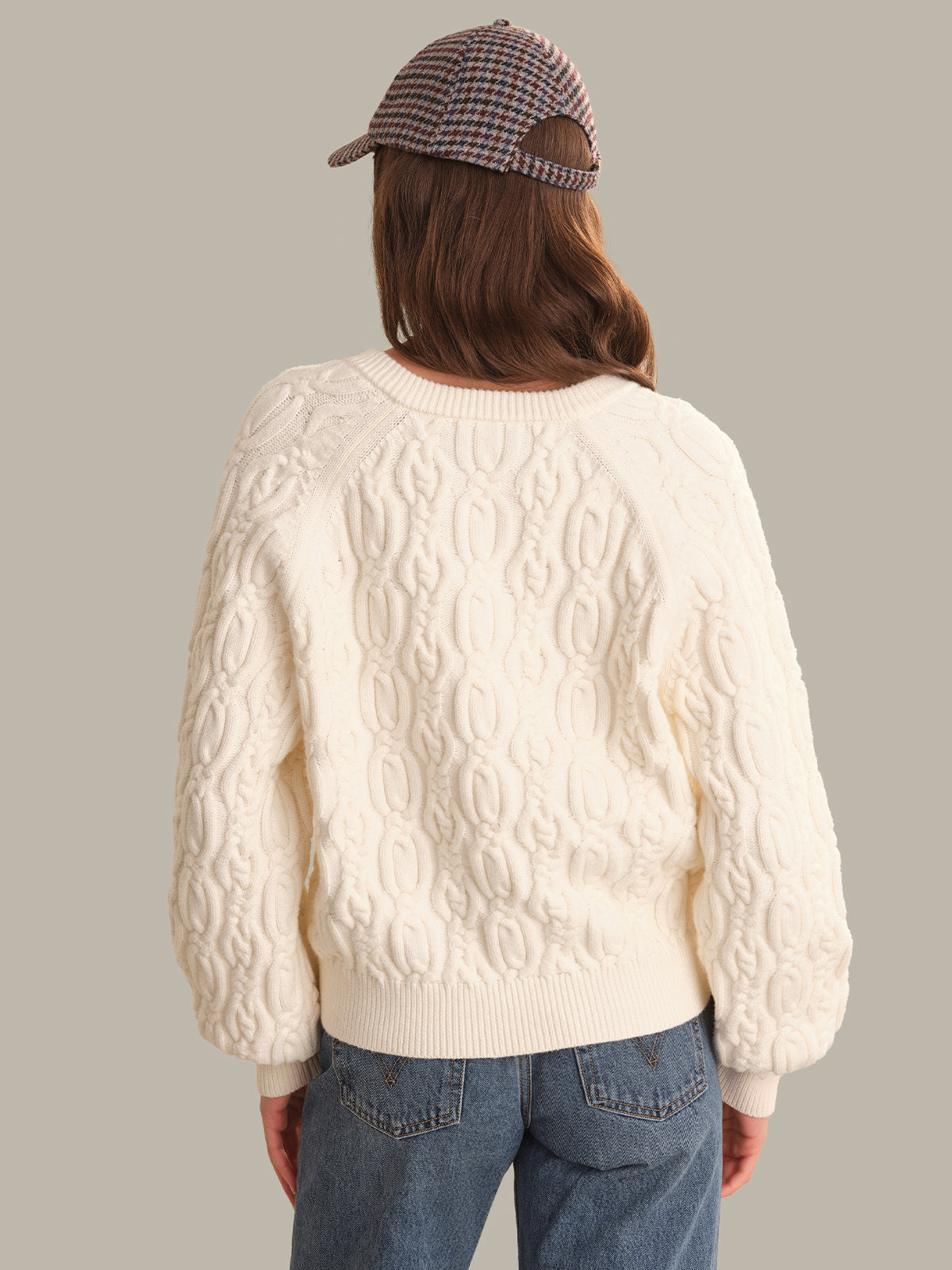 A person shown from the back wearing a cream cable-knit sweater, blue jeans, and a brown and beige plaid baseball cap, against a solid light gray background.