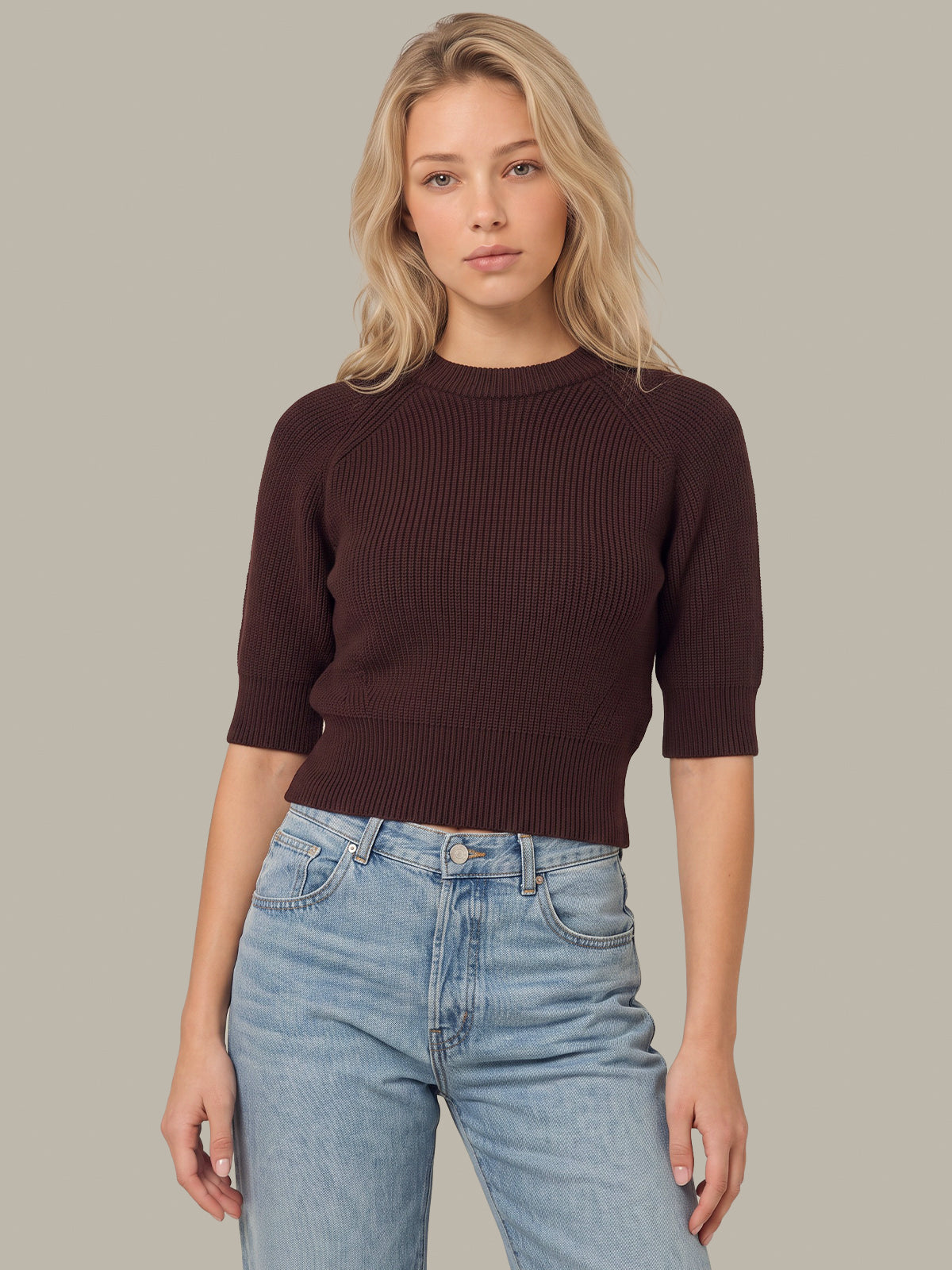 A woman modeling a dark brown ribbed knit sweater with three-quarter sleeves and a crew neck, paired with light-wash blue jeans, against a solid taupe background.