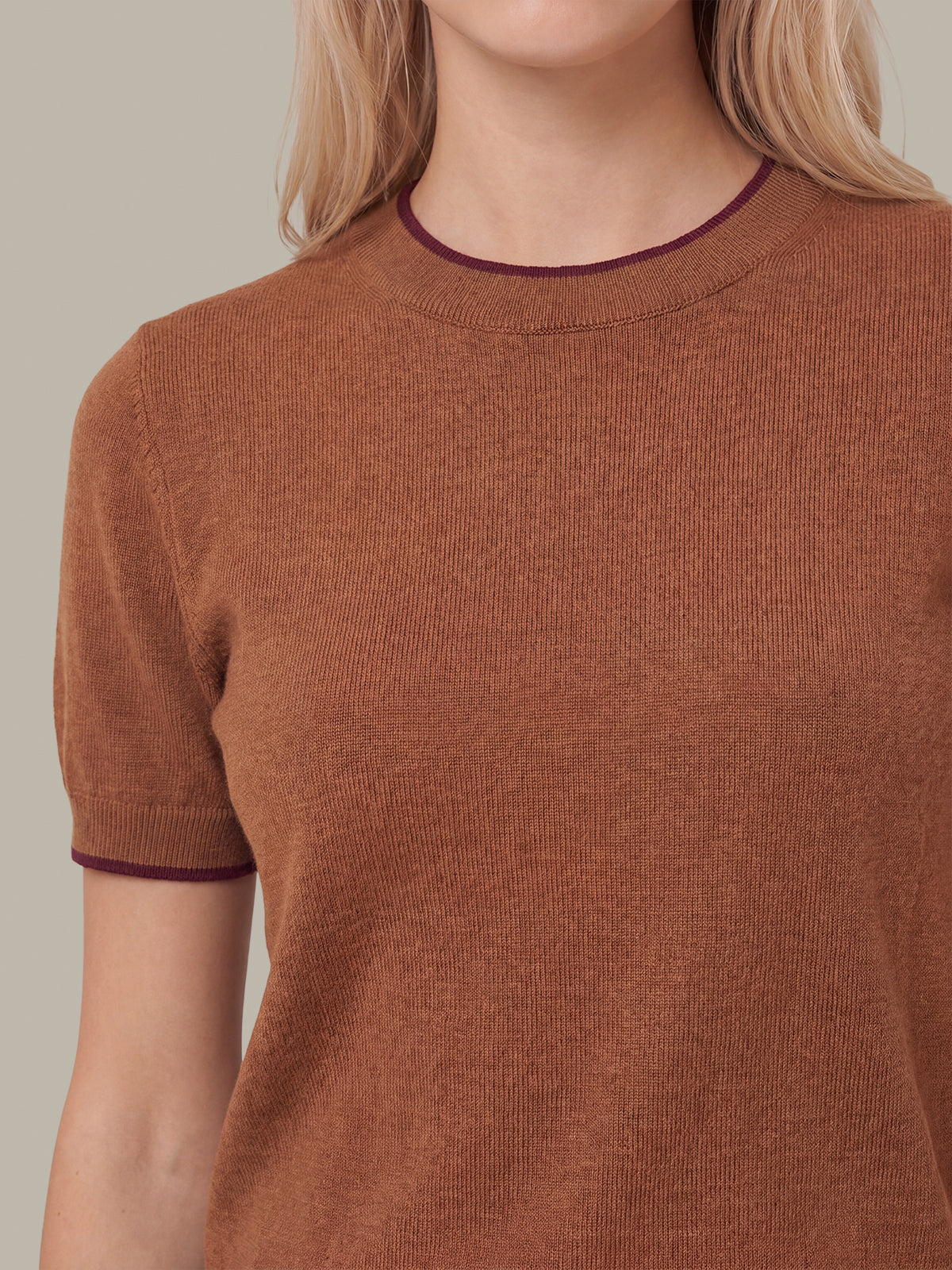 A close-up of a woman wearing a short-sleeve ribbed knit sweater in a warm rust brown color, featuring contrasting dark burgundy trim at the crew neck and sleeve cuffs.