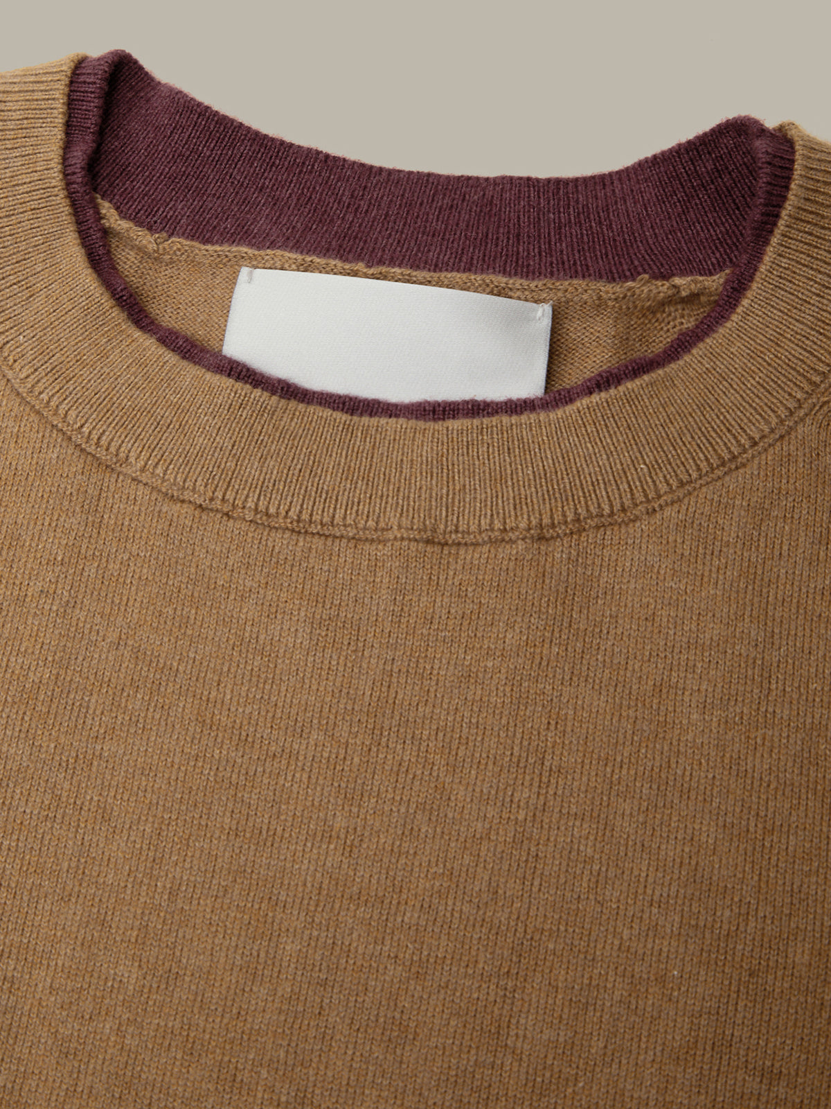 Close-up of a ribbed knit crewneck sweater with a warm camel brown body and a contrasting dark plum purple inner collar, featuring a blank white rectangular label.