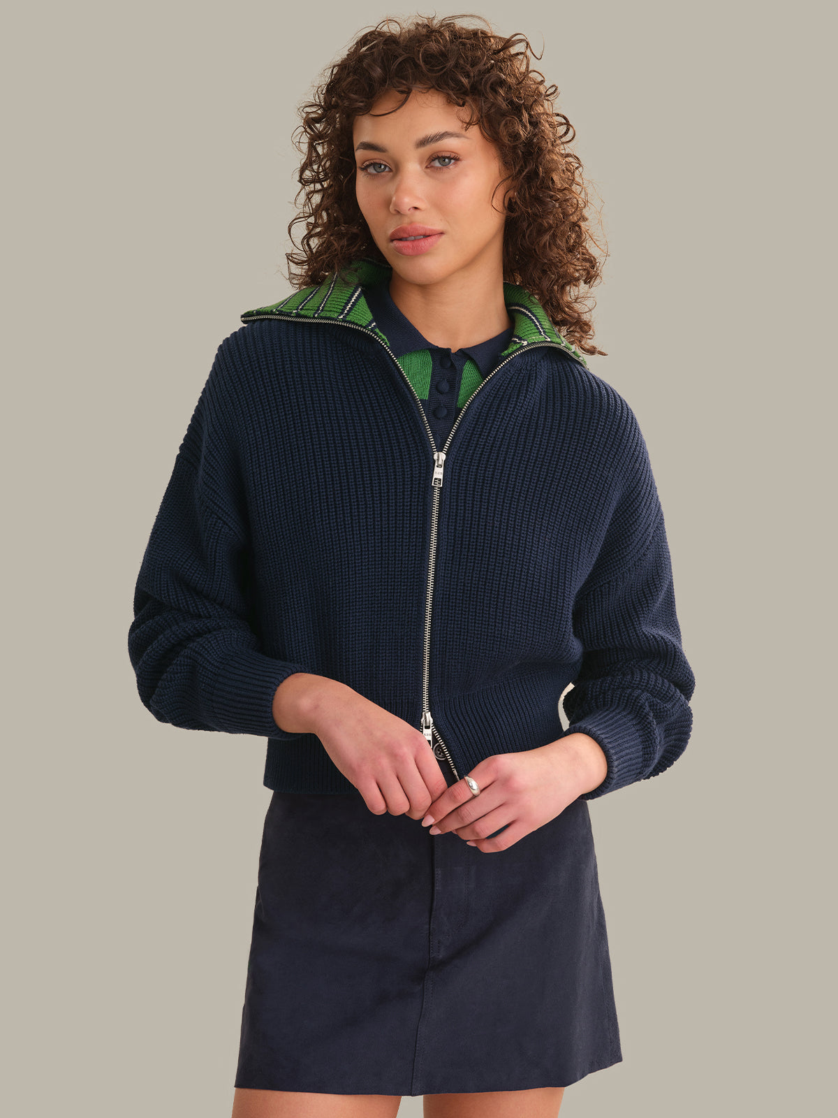 A model with curly brown hair wearing a navy blue ribbed zip-up hoodie with a green patterned inner hood lining and a matching navy mini skirt, set against a solid gray background.