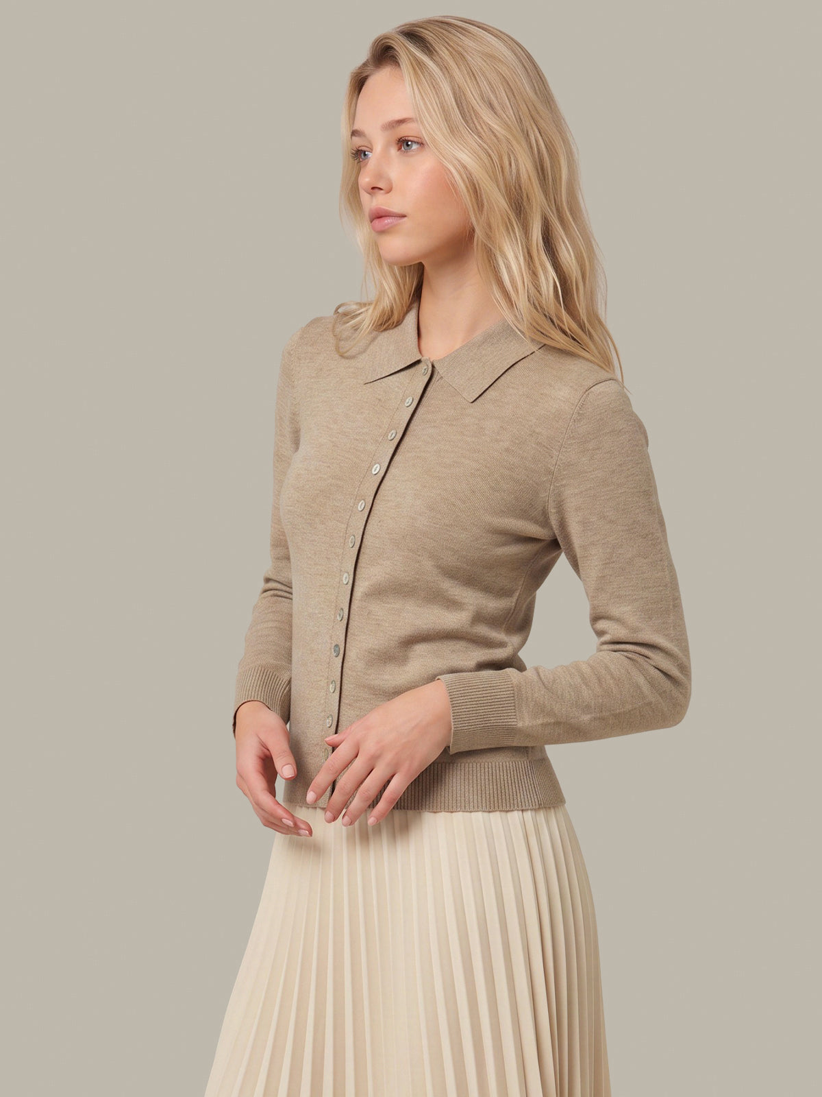 A woman wearing a beige knit button-front cardigan with a collar and a cream pleated skirt, posed against a solid light gray studio background.