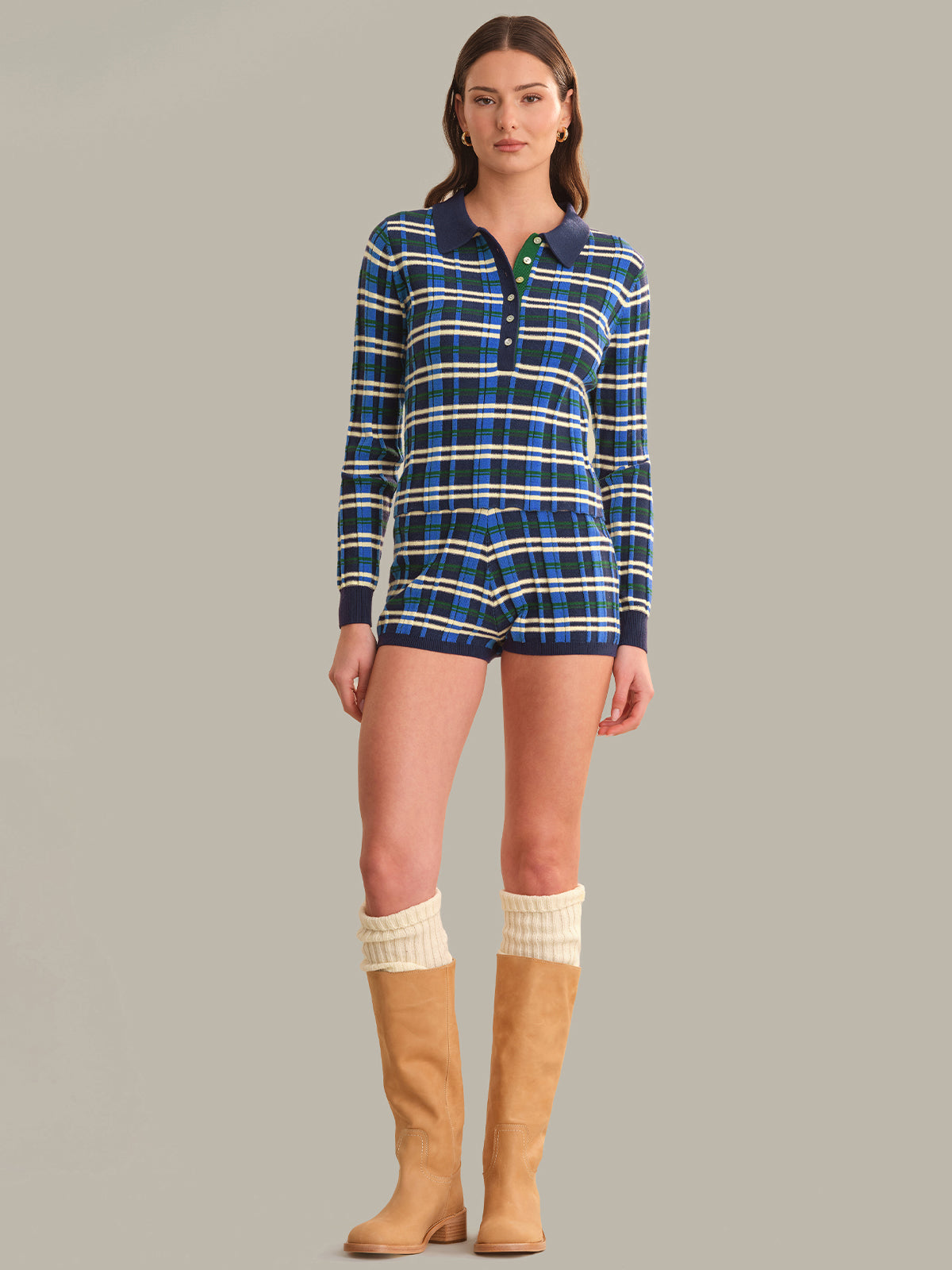A model wearing a matching long-sleeve plaid polo shirt and shorts set in blue, white, and yellow, paired with cream leg warmers and tan knee-high boots, against a solid gray background.