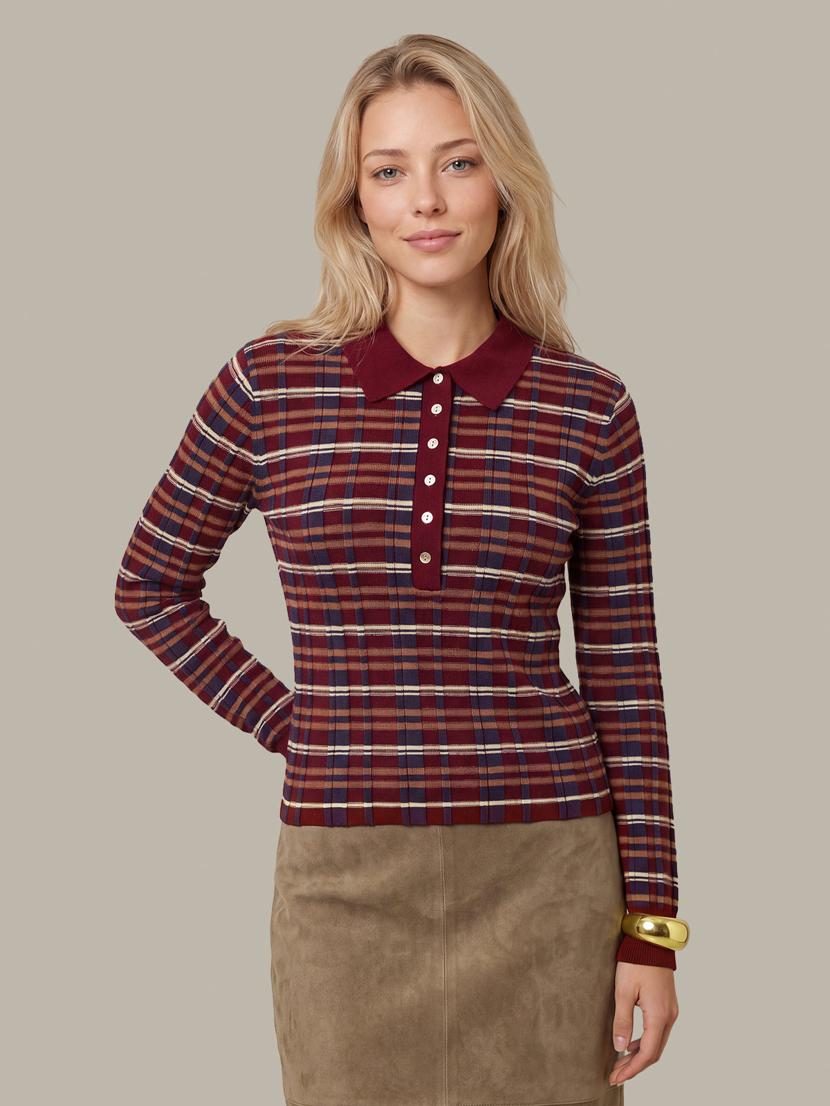A woman modeling a long-sleeve knit polo shirt with horizontal red, navy, and white plaid stripes, featuring a solid burgundy collar and button placket, paired with a brown skirt against a solid beige background.