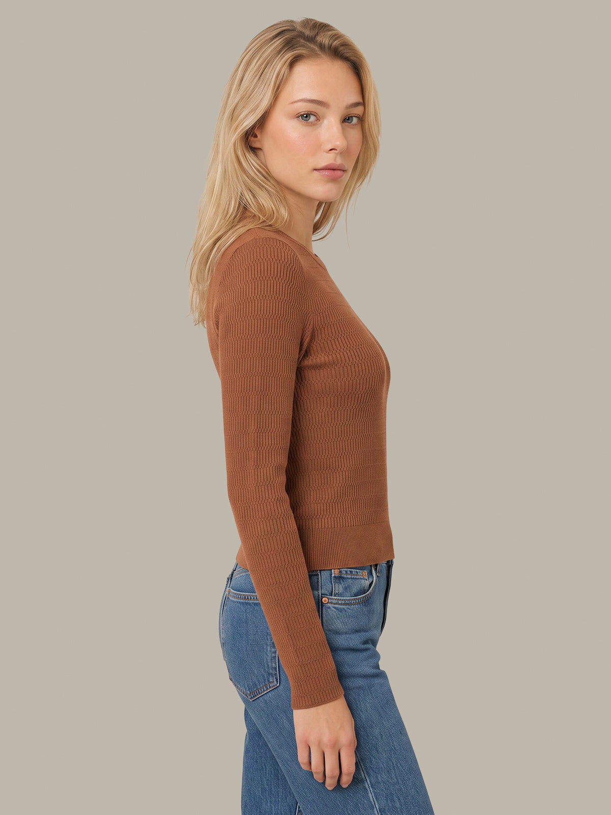 A woman shown in side profile wearing a fitted ribbed long-sleeve knit top in a warm camel brown color, paired with blue jeans, against a solid light gray background.