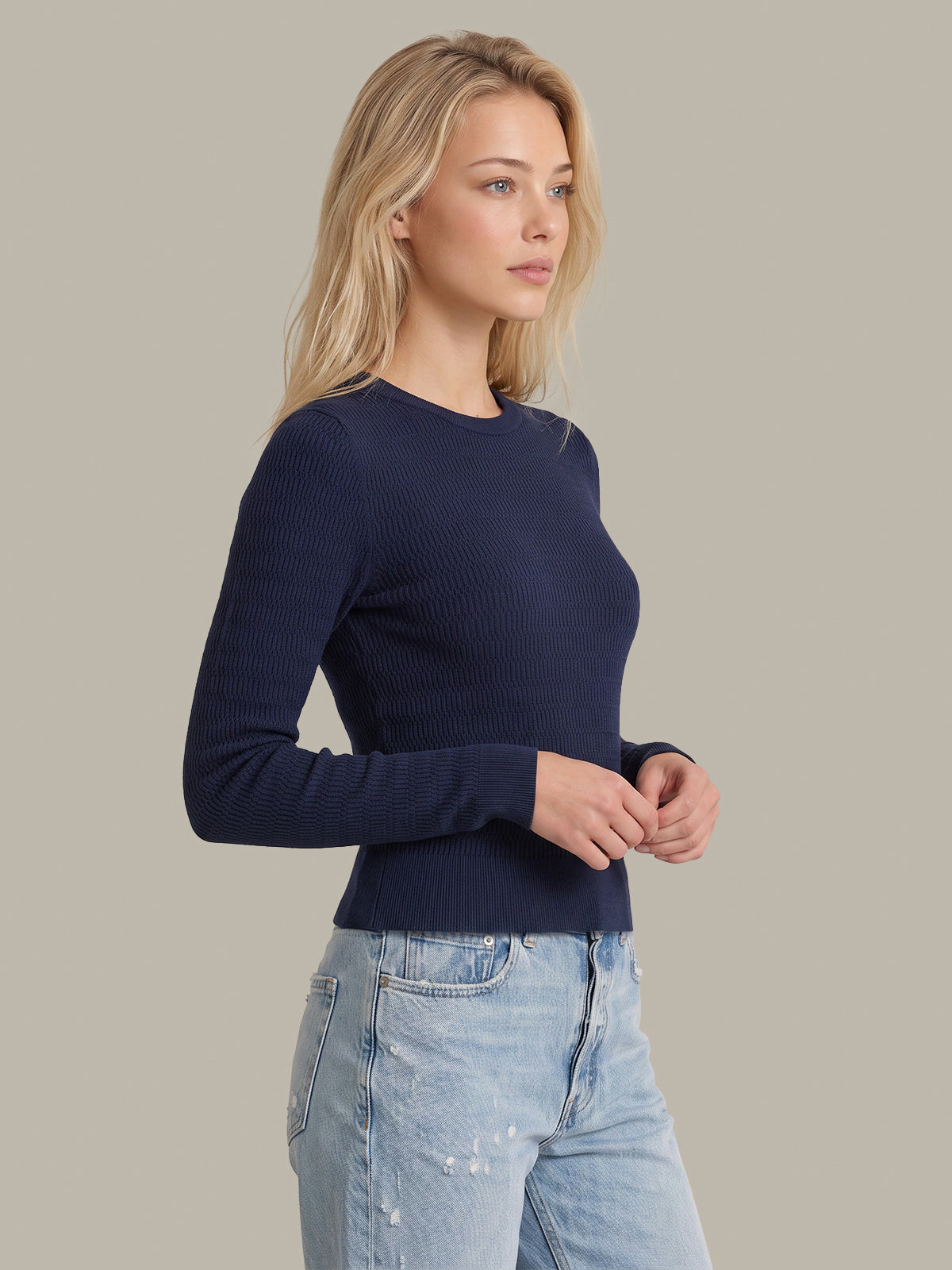 A woman wearing a navy blue ribbed long-sleeve crewneck sweater and light wash blue jeans, standing against a solid light gray studio background.