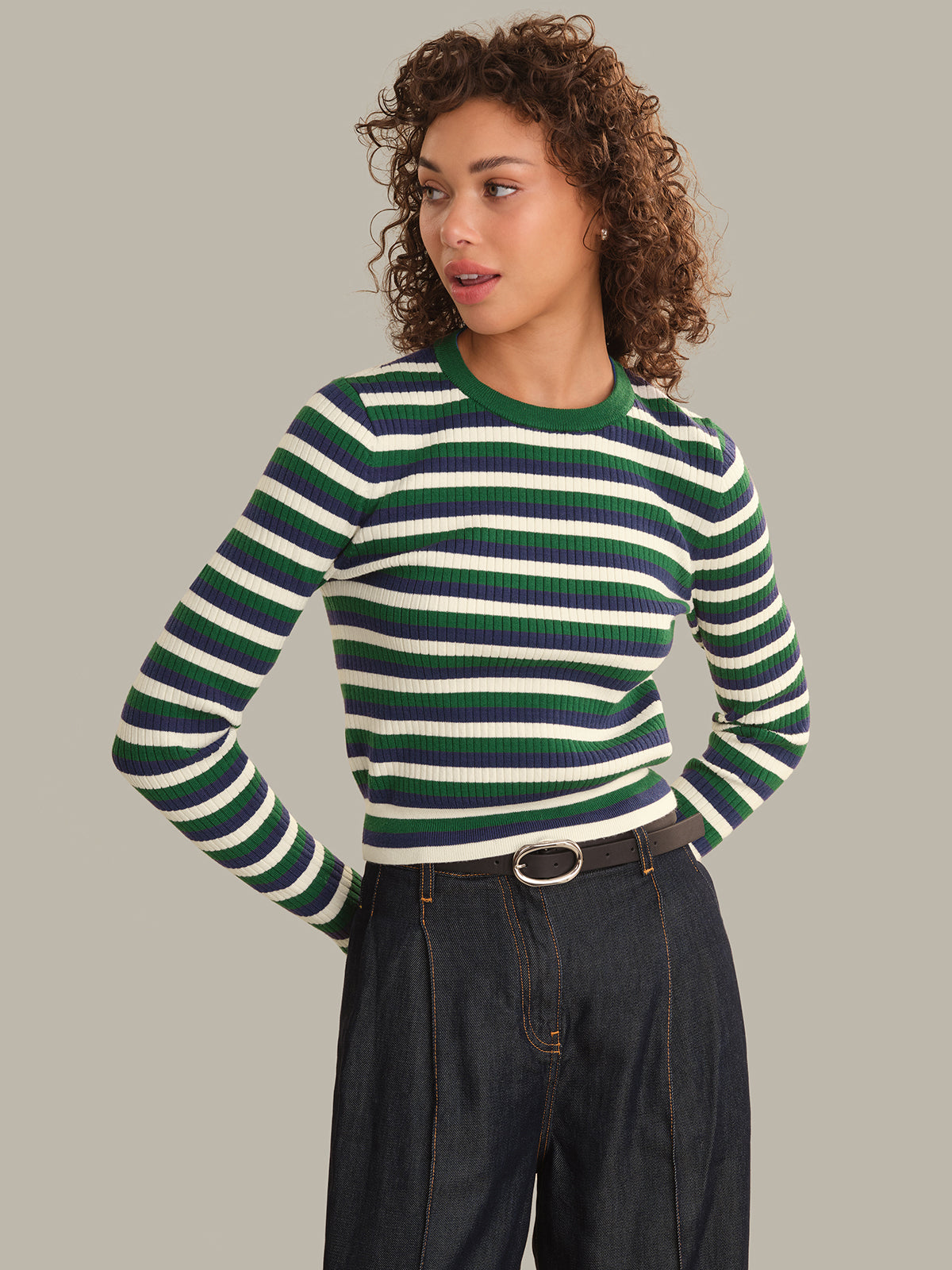 A woman wearing a green and white striped ribbed knit sweater with a dark waist belt and dark jeans, standing against a solid light gray background.