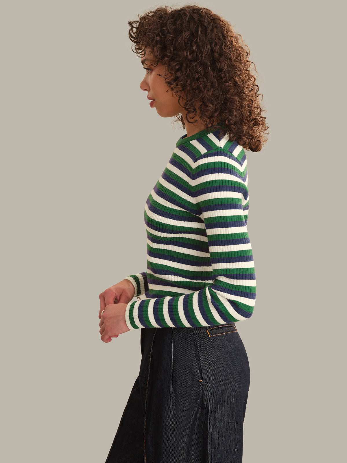 A woman with curly brown hair is shown in profile wearing a fitted long-sleeve knit sweater with horizontal green, navy, and white stripes, paired with dark denim bottoms, against a solid light gray background.