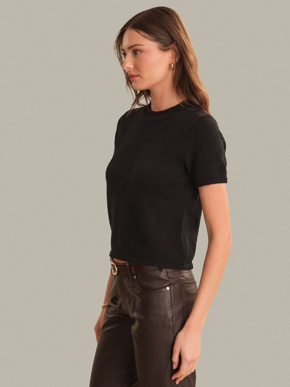A woman wearing a black short-sleeve ribbed knit top and dark brown leather pants, shown in a side-profile studio shot against a solid light gray background.