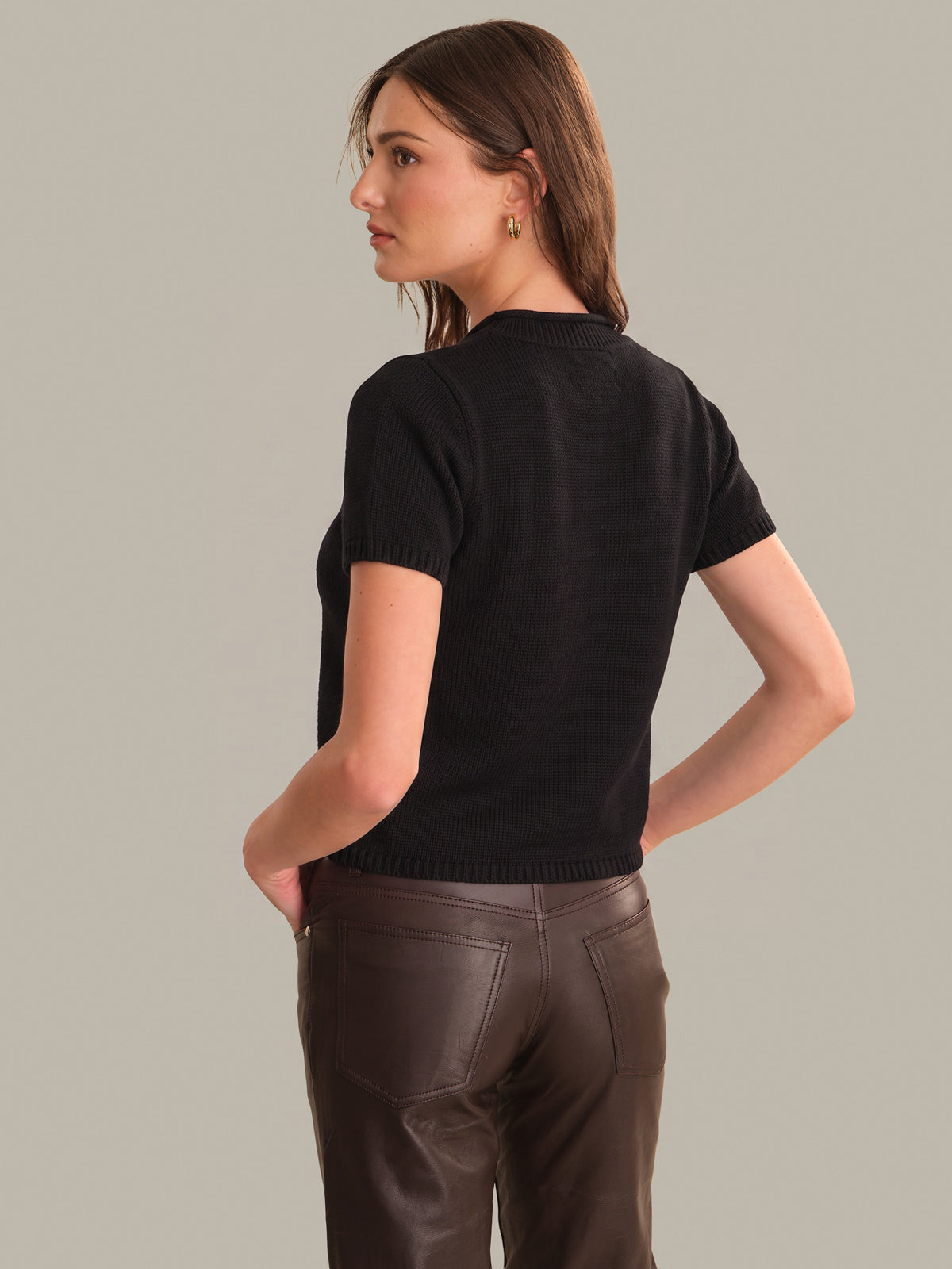 A woman shown from the back wearing a black short-sleeve knit top and dark brown pants against a plain light gray studio background.