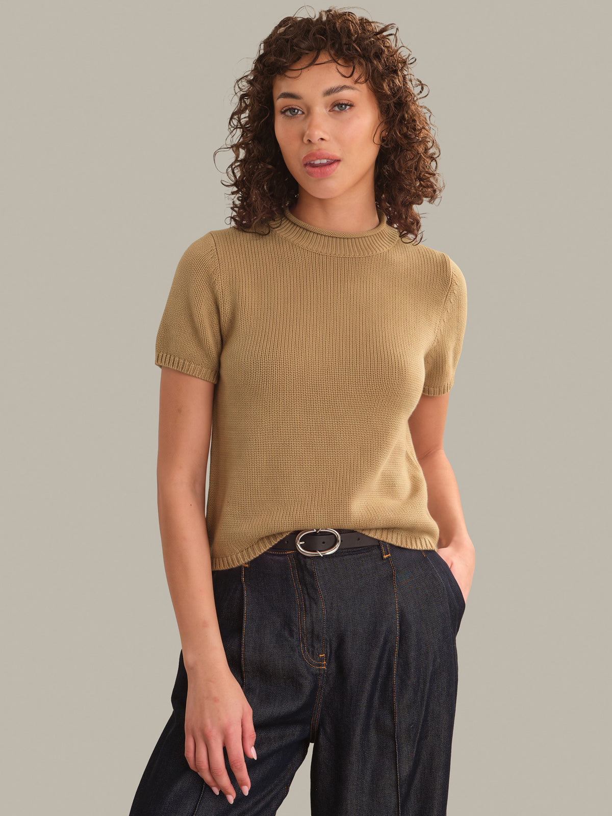 A woman with curly brown hair wearing a short-sleeve ribbed knit sweater in camel beige and dark wide-leg jeans, standing against a solid light gray studio background.