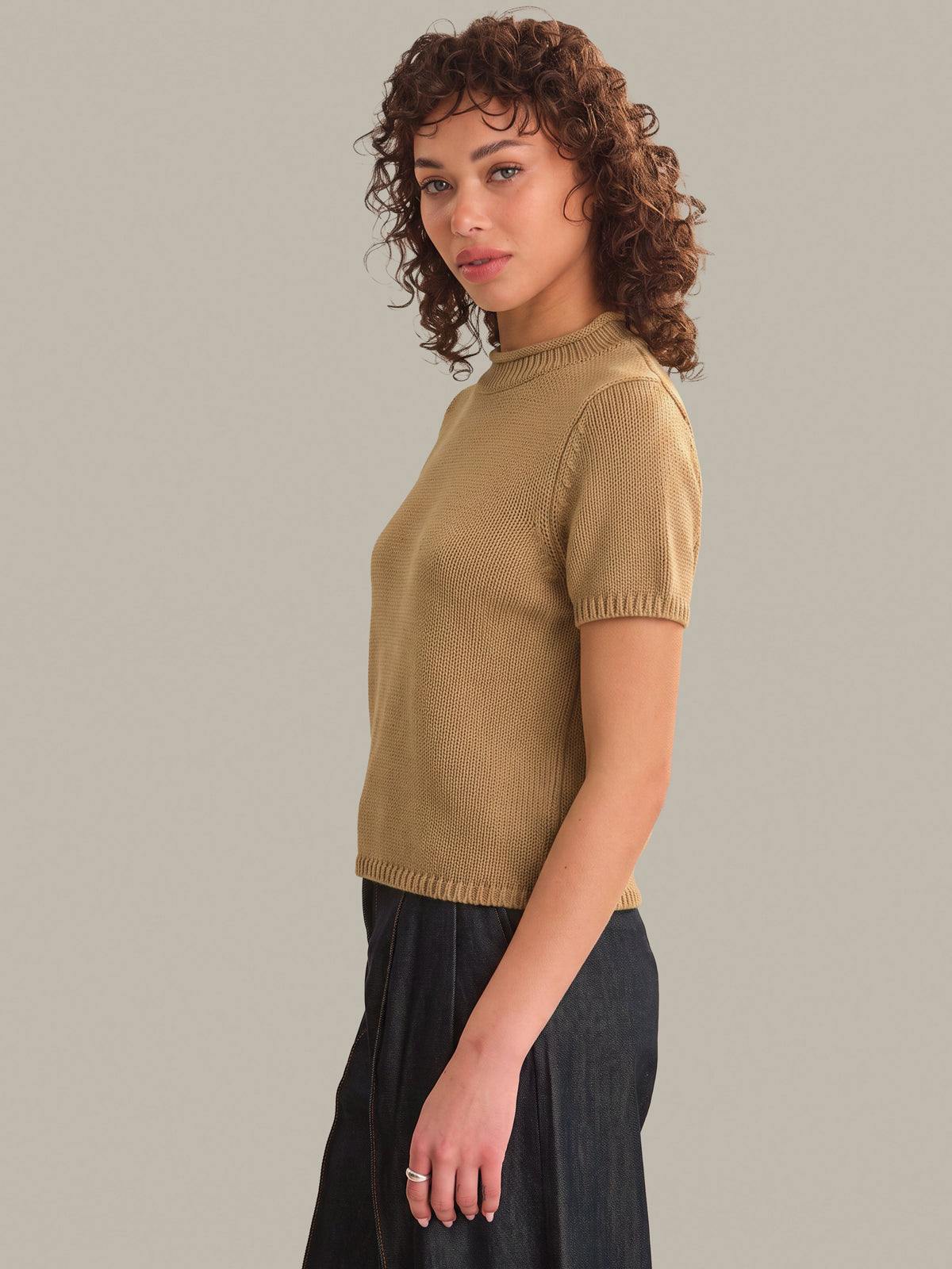 A woman with curly brown hair wearing a short-sleeve ribbed knit top in camel tan and dark wide-leg pants, photographed against a solid light gray studio background.