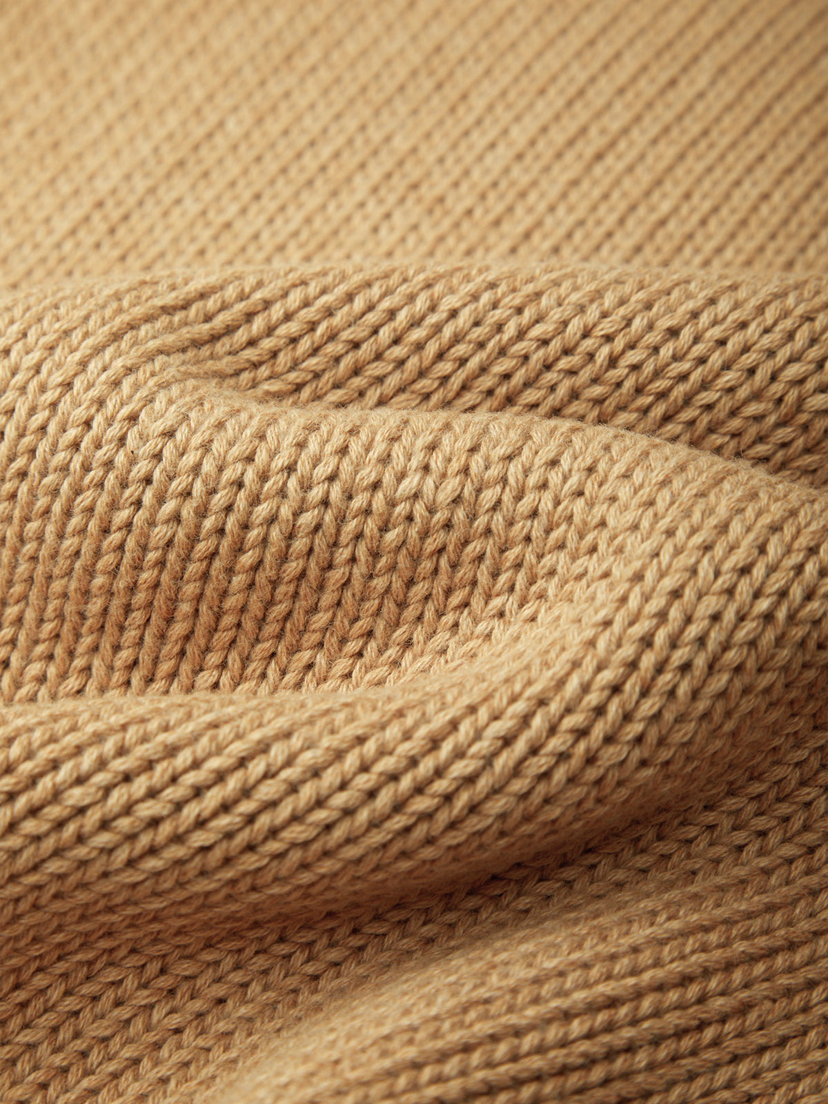 Close-up of a soft knit fabric in a warm camel beige color, showing the detailed texture and folds of the yarn.