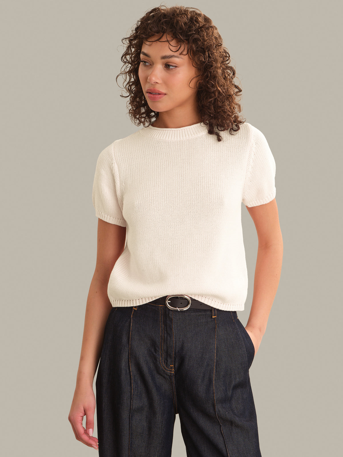 A woman with curly brown hair wearing a short-sleeve ribbed cream knit sweater and dark wide-leg jeans with a silver-belt, photographed against a solid light gray studio background.