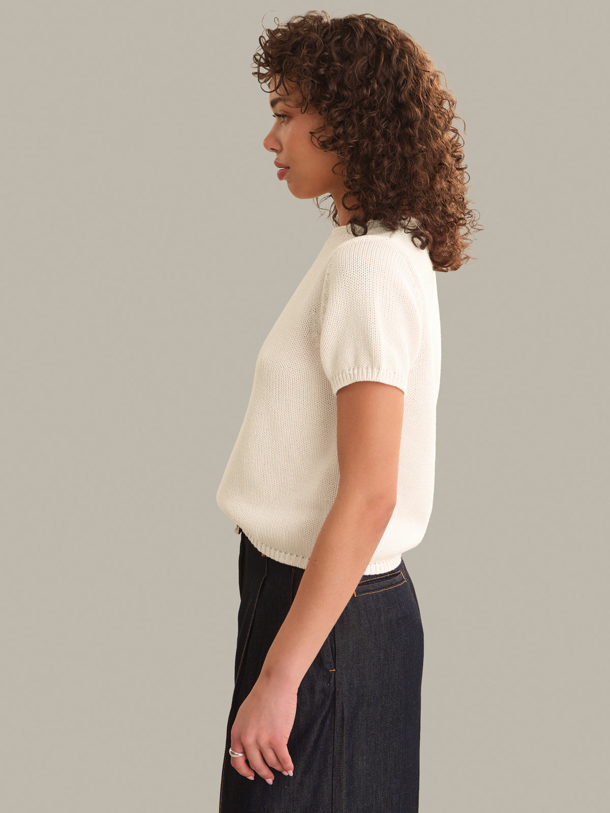 A woman with curly brown hair is shown in profile wearing a cream-colored short-sleeve ribbed knit top and dark denim jeans, set against a solid light gray studio background.