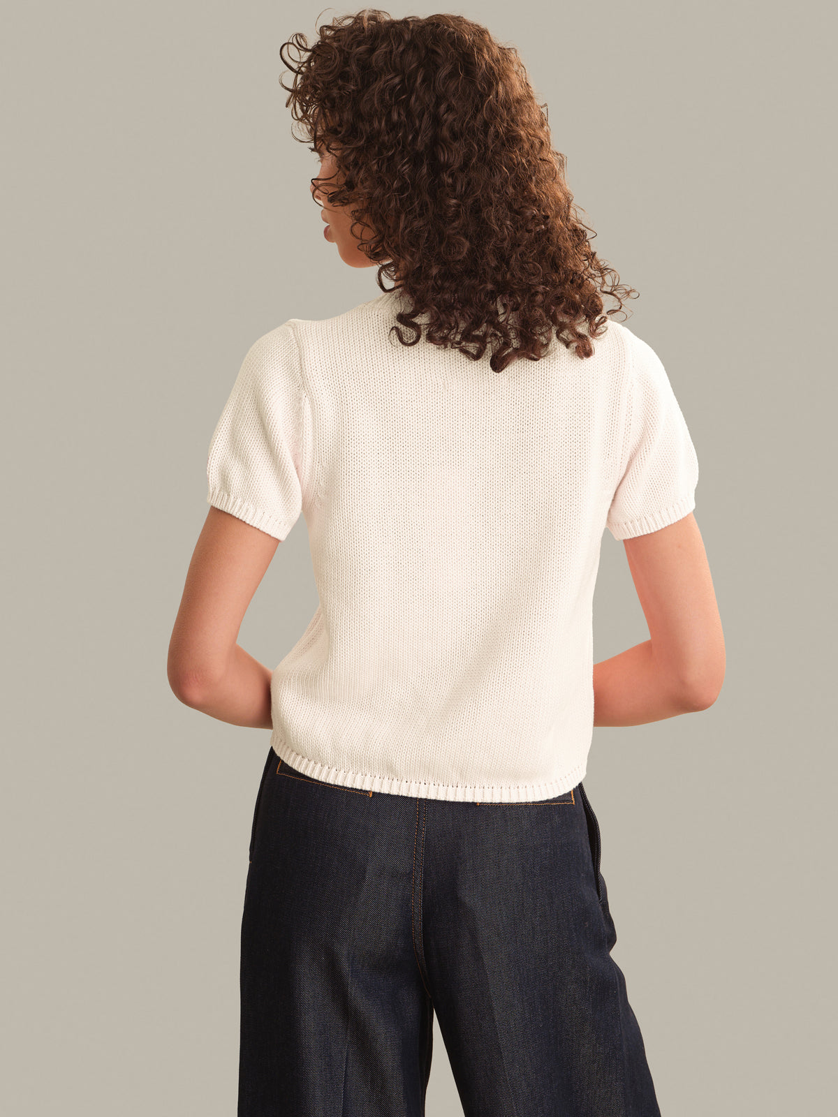 A woman with curly brown hair is shown from behind wearing a short-sleeve cream knit top and dark blue jeans, set against a solid light gray background.