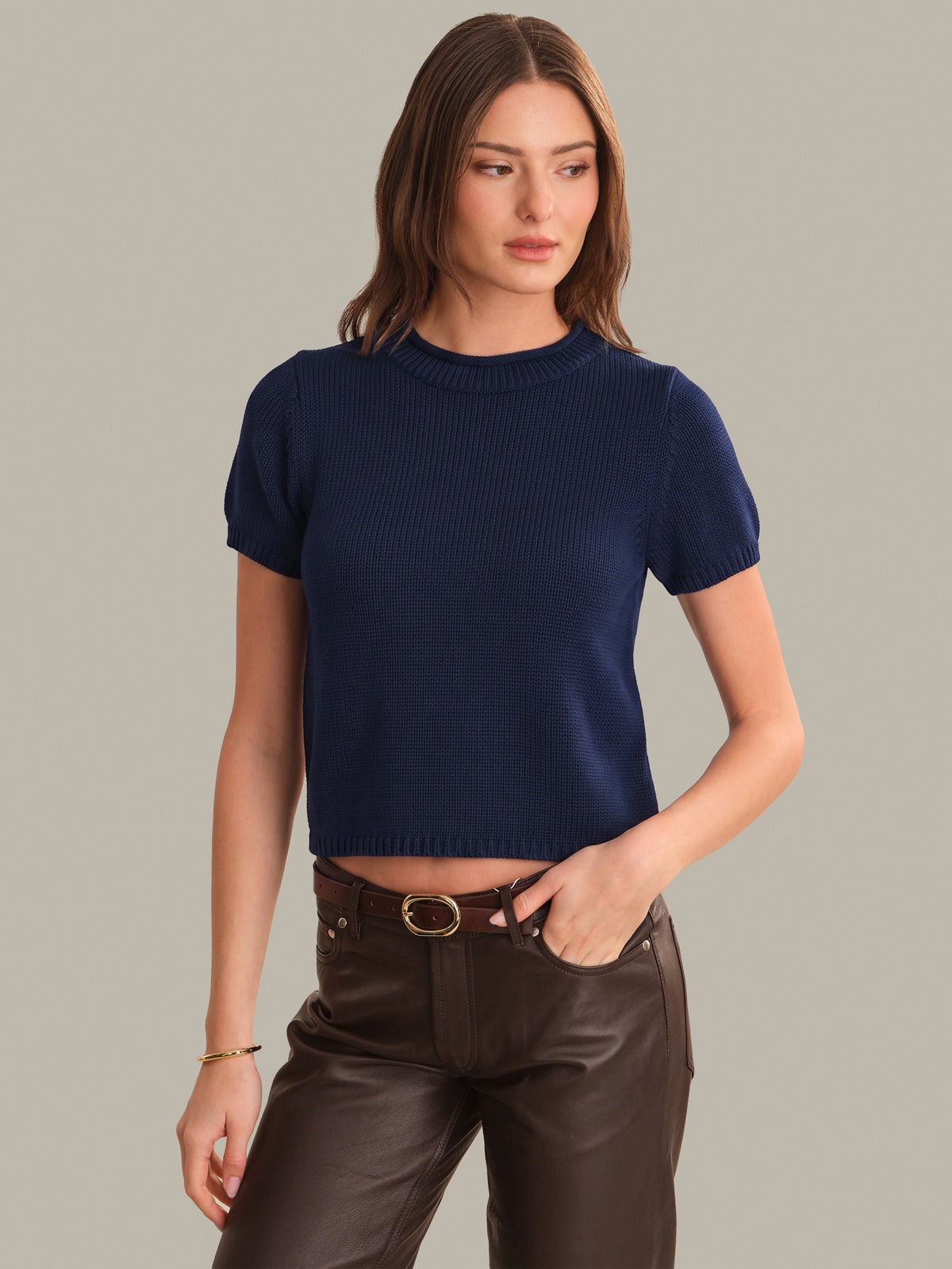 A woman modeling a navy blue ribbed short-sleeve crop top paired with dark brown pants, against a solid light gray background.