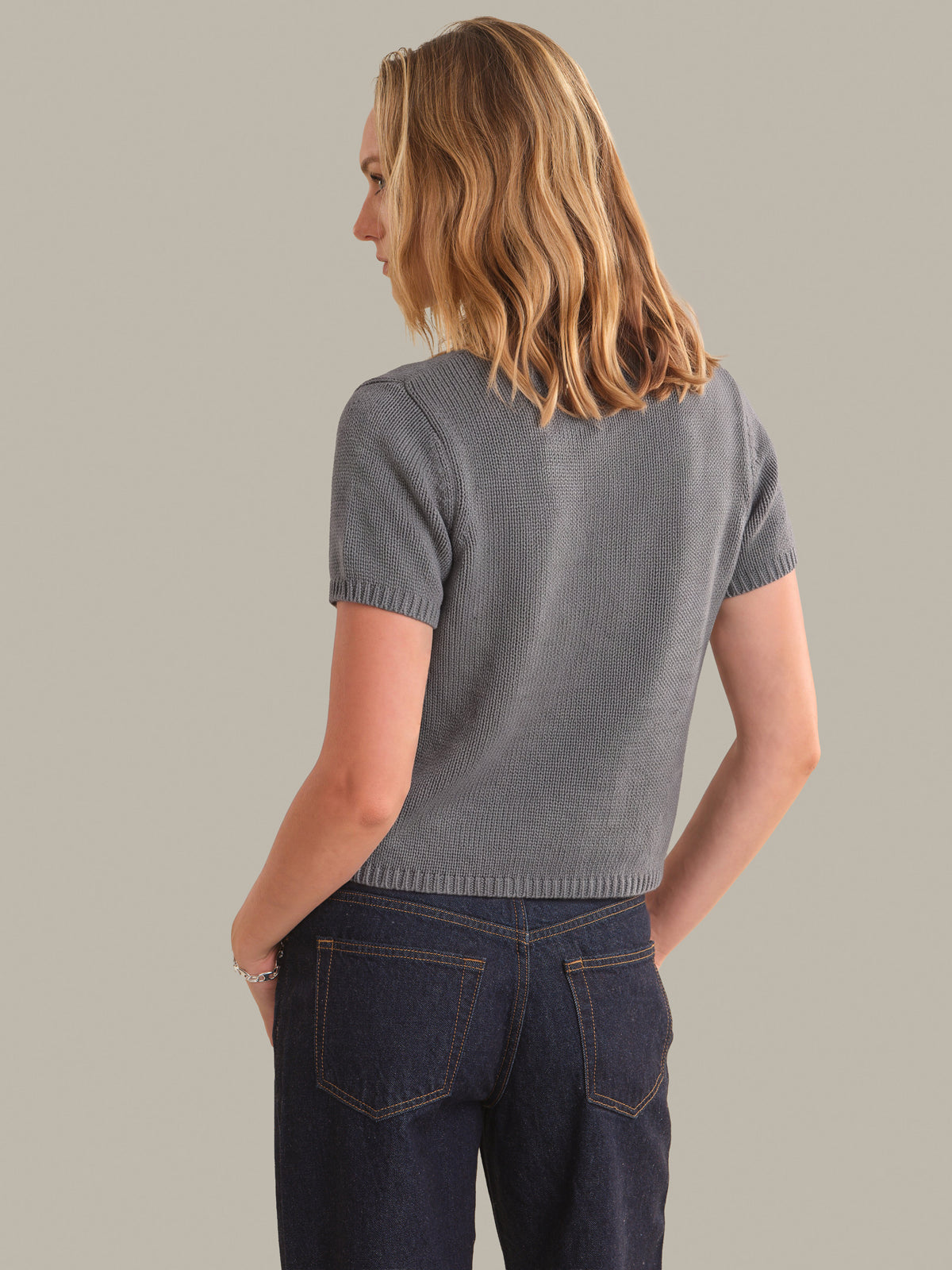 A woman shown from the back wearing a short-sleeve ribbed knit top in a heather gray color, paired with dark blue jeans, against a solid light gray background.