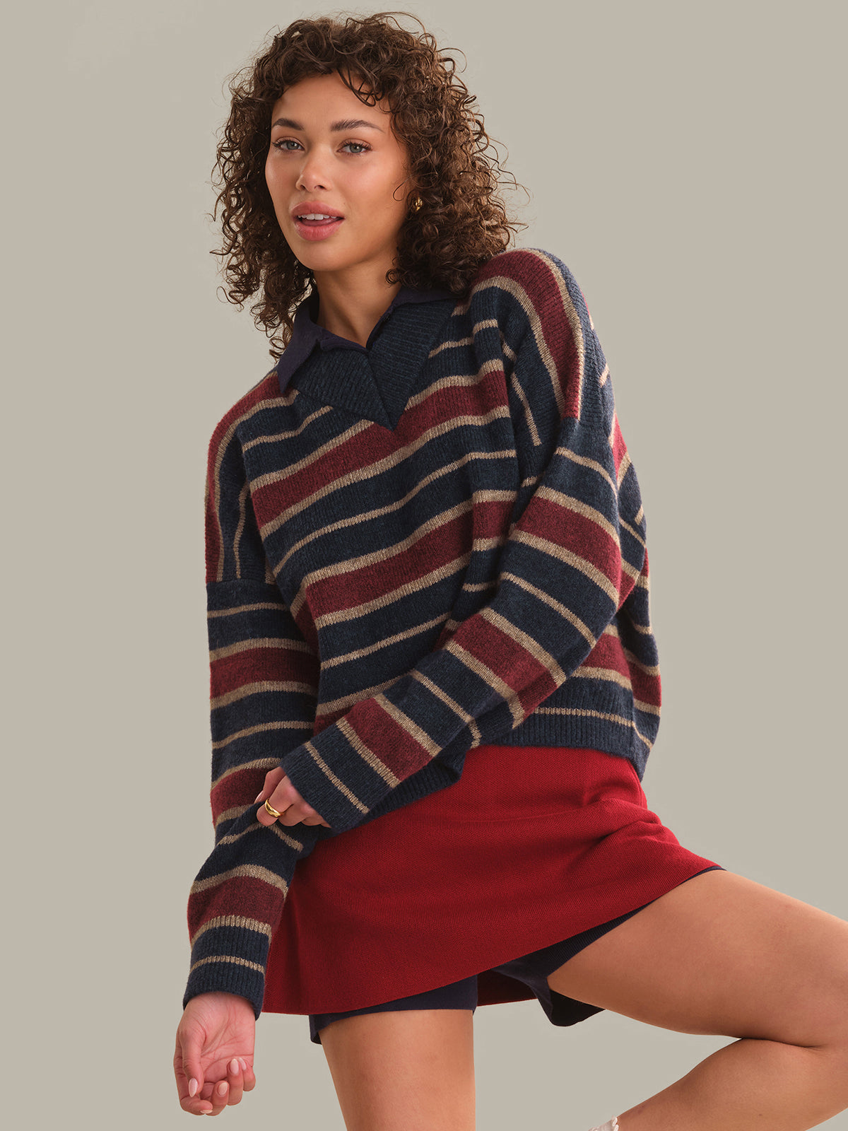 A woman modeling a striped knit sweater with a polo collar, featuring navy, burgundy, and beige horizontal stripes, paired with a red mini skirt against a solid beige background.