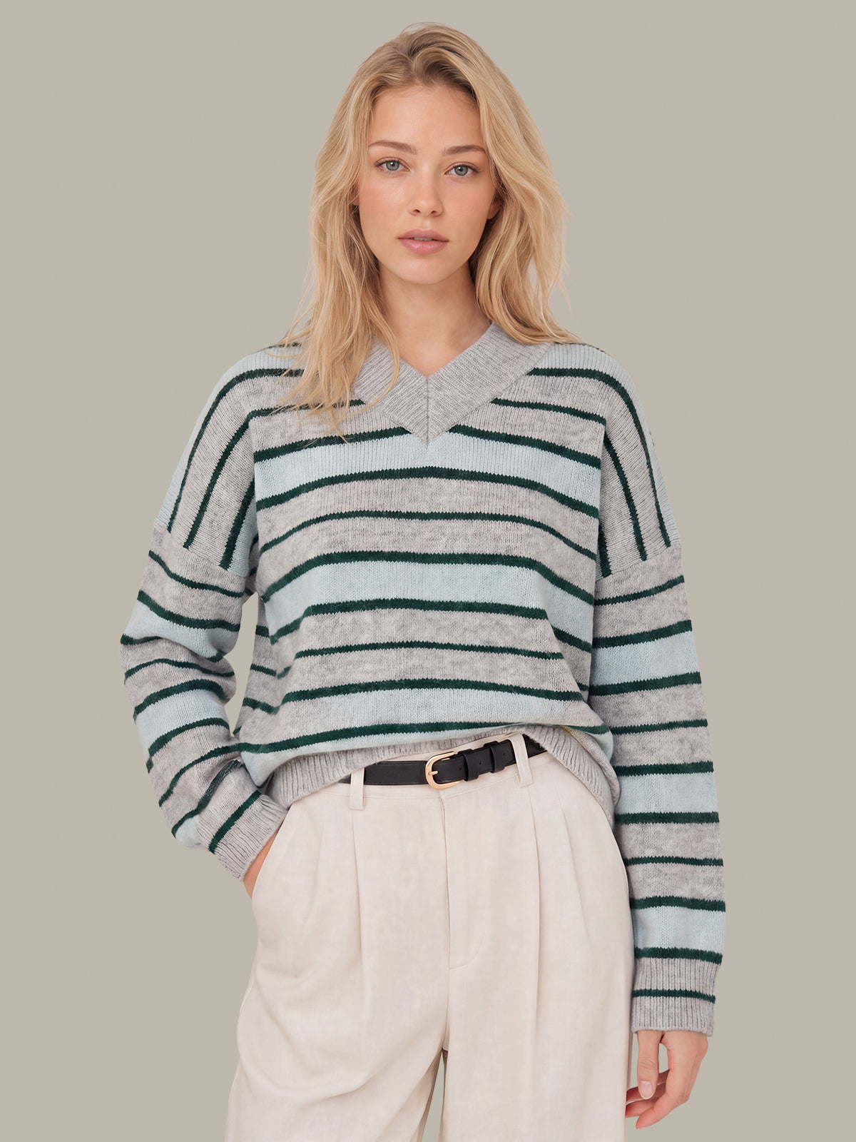A woman modeling a gray and light blue striped knit sweater with dark green stripes, paired with cream trousers, against a solid taupe background.