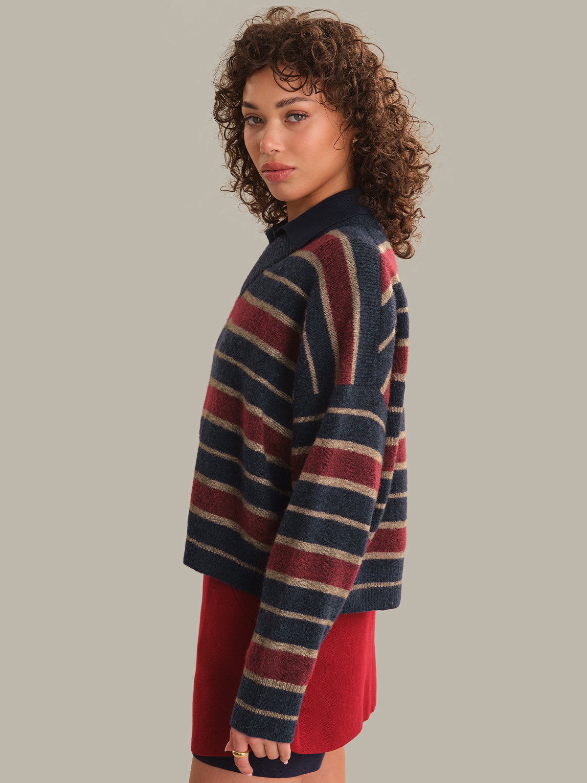 A woman with curly brown hair is shown in profile wearing a cropped, long-sleeve knit sweater with horizontal stripes in navy blue, burgundy red, and cream, paired with a solid red skirt, against a solid light gray background.