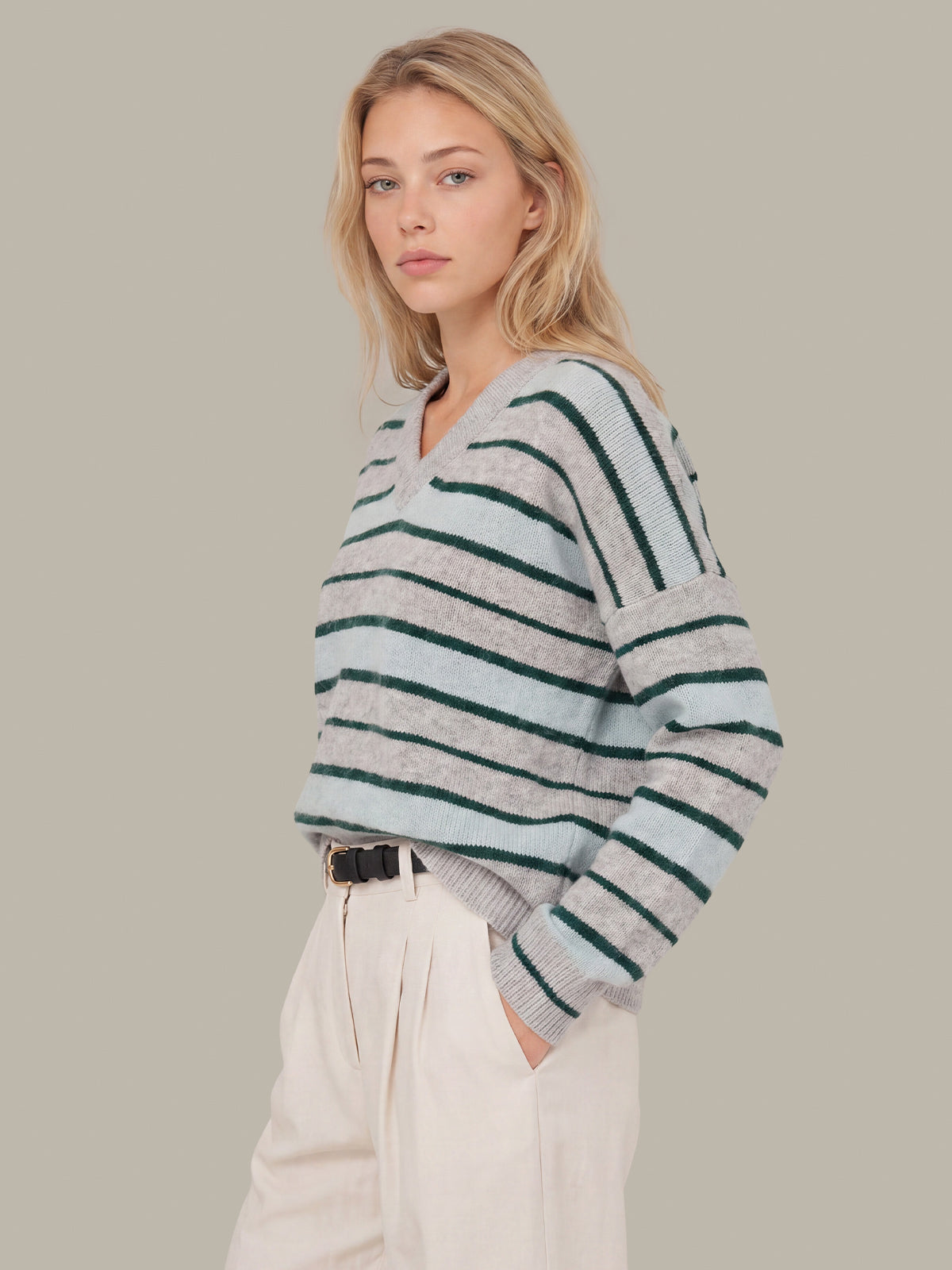 A woman wearing a light gray and dark green striped knit sweater with a V-neck, paired with cream trousers, against a solid beige background.