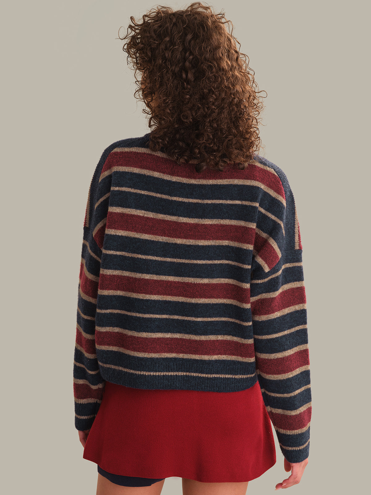 A person shown from the back wearing a cropped knit sweater with horizontal navy, burgundy, and cream stripes, paired with a red skirt, against a plain warm gray background.
