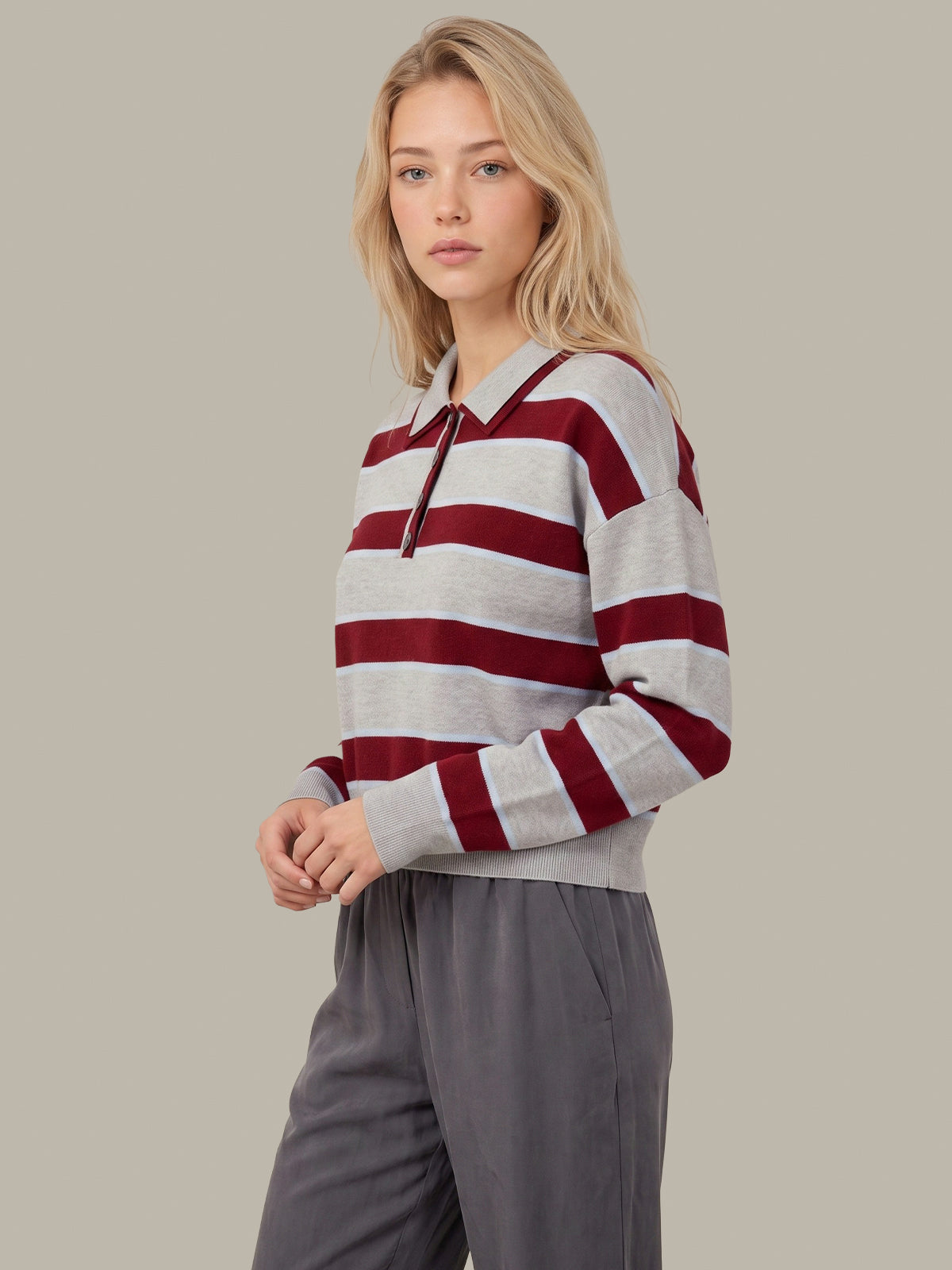 A woman modeling a long-sleeve knit polo-style top with horizontal burgundy and light gray stripes, paired with gray pants, against a plain beige studio background.