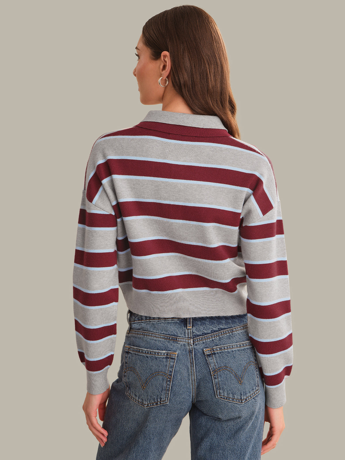 A woman shown from the back wearing a cropped long-sleeve knit sweater with horizontal burgundy and light gray stripes, paired with blue jeans, against a solid light gray background.