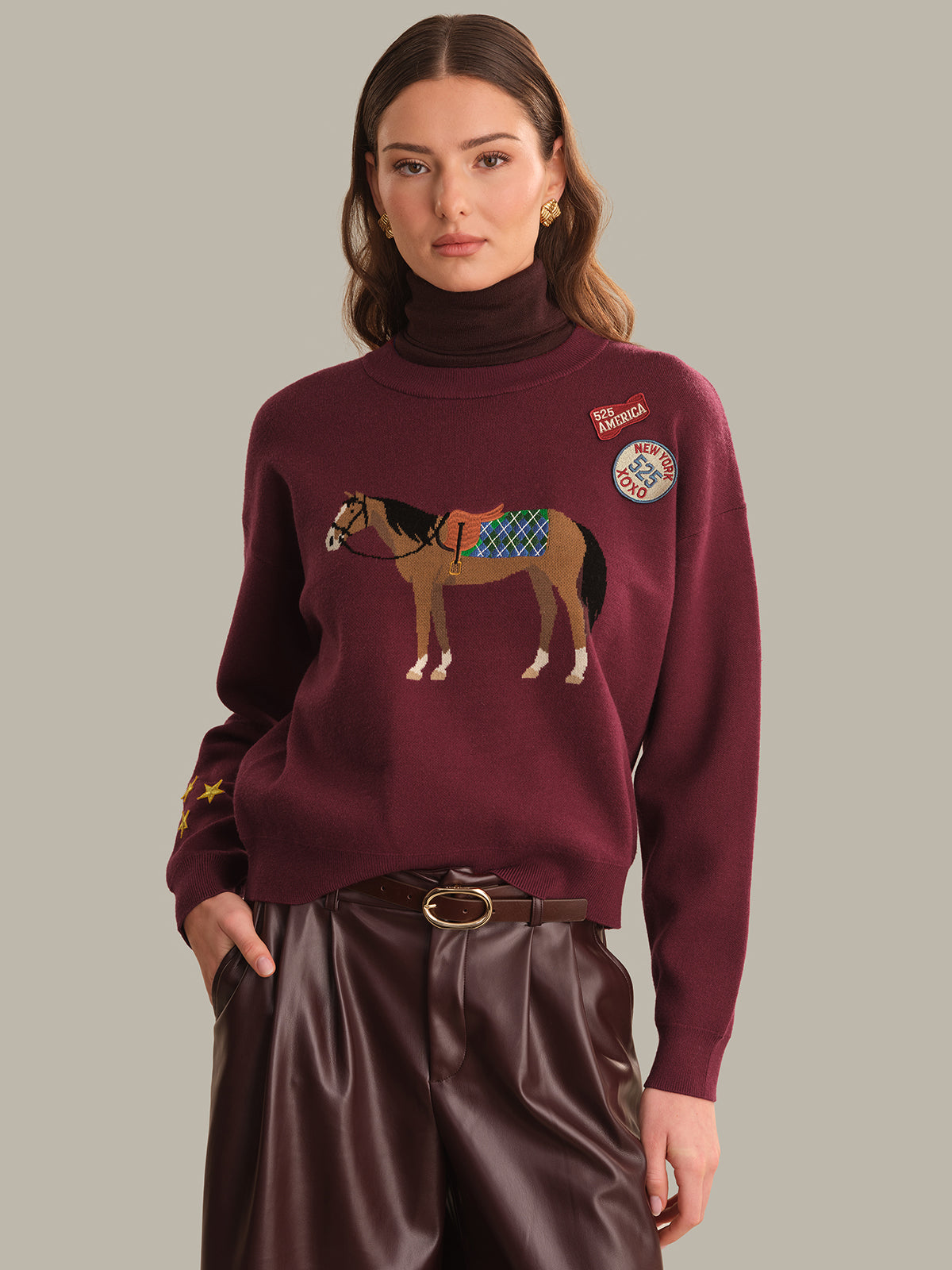 A woman wearing a burgundy crewneck sweater with a horse graphic and embroidered patches, paired with dark brown pants, against a solid gray background.