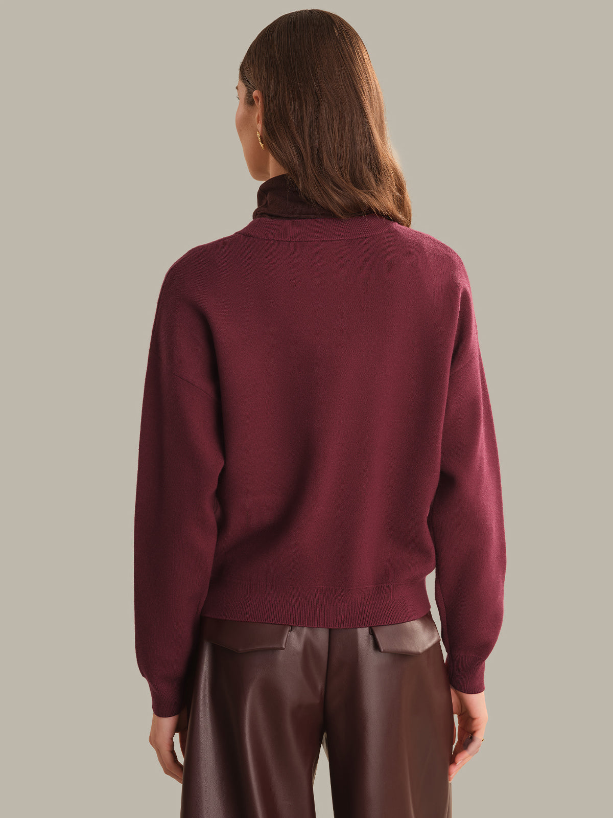 A woman shown from the back wearing a burgundy crewneck knit sweater and dark brown pants, set against a solid light gray studio background.