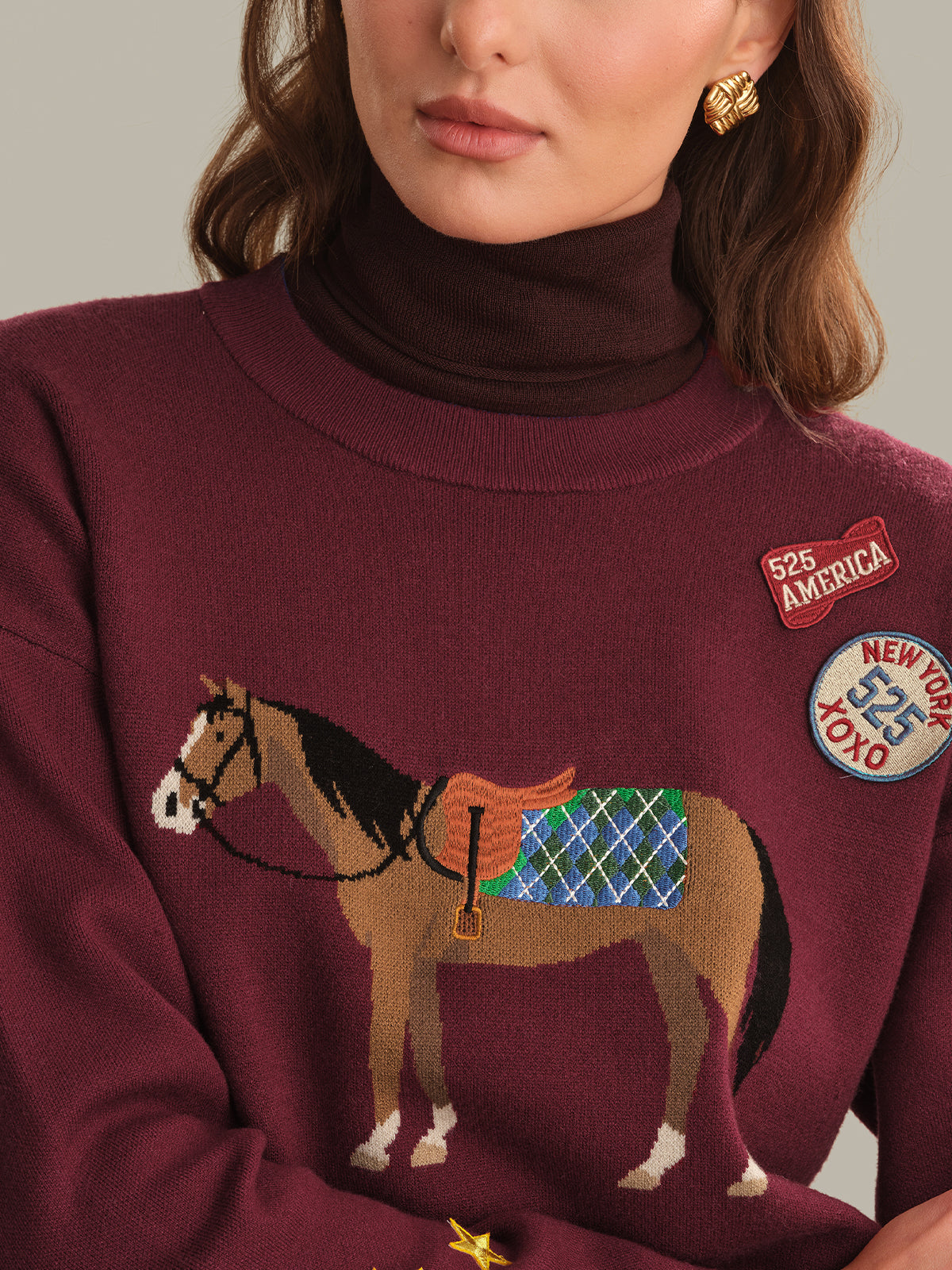 A close-up of a woman wearing a burgundy knit sweater with a large embroidered horse design and decorative patches on the chest, paired with a dark brown turtleneck underneath.