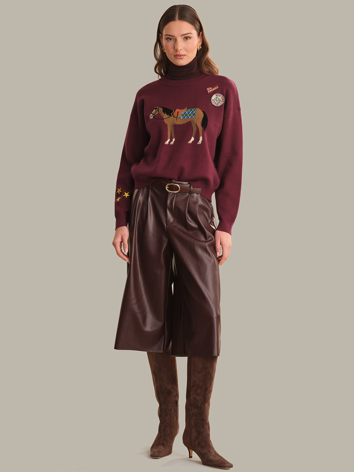 A model stands against a solid gray background wearing a burgundy crewneck sweater with a horse graphic and patches, paired with dark brown wide-leg leather pants and matching brown knee-high boots.