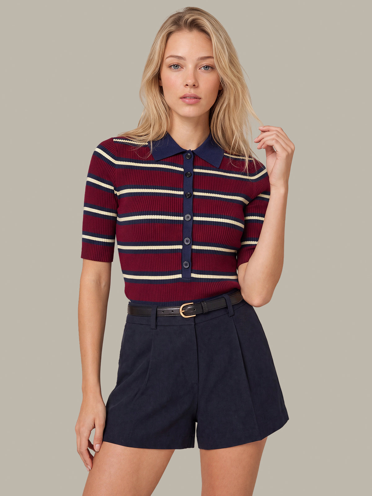 A blonde model wearing a short-sleeve ribbed knit polo shirt with navy blue and cream horizontal stripes, paired with dark navy shorts, against a solid taupe background.