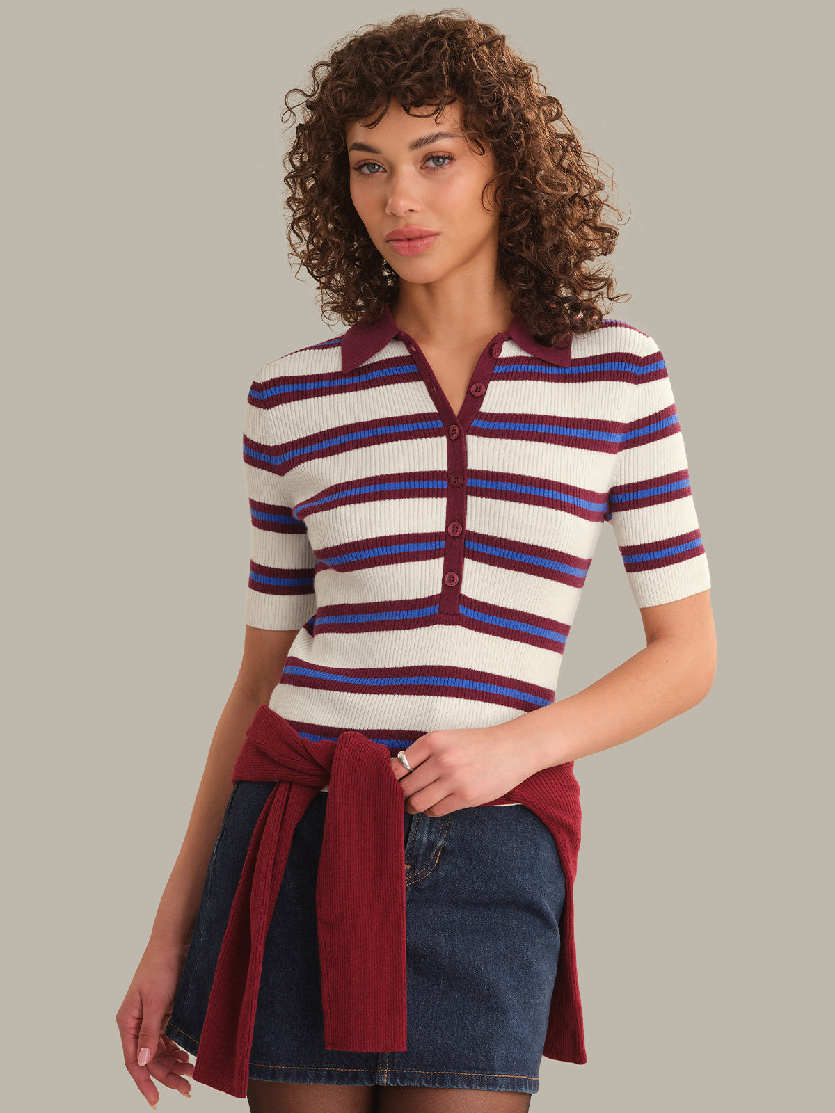 A woman with curly brown hair wearing a short-sleeve ribbed knit polo shirt with horizontal cream, blue, and burgundy stripes, paired with a dark denim skirt and a tied burgundy sweater, against a solid taupe background.