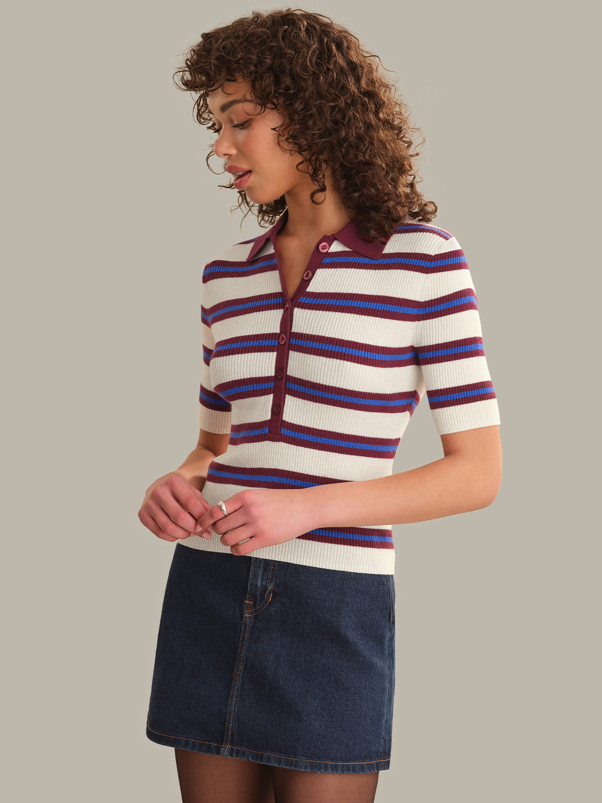 A woman wearing a short-sleeve knit polo-style top with horizontal cream, burgundy, and blue stripes, paired with a dark denim mini skirt, against a plain warm gray studio background.