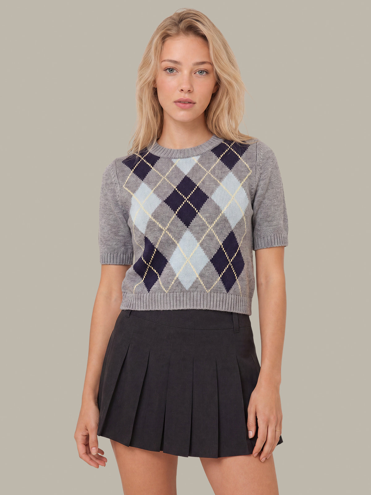 A woman modeling a short-sleeve knit sweater with an argyle pattern in gray, navy, light blue, and beige, paired with a dark pleated mini skirt, against a solid taupe background.