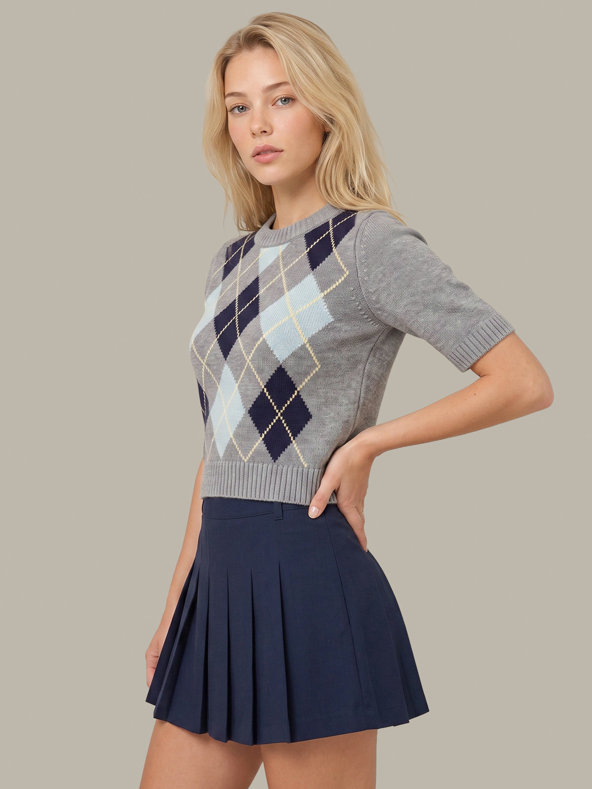 A blonde model wearing a gray short-sleeve knit sweater with a navy, light blue, and beige argyle pattern on the chest, paired with a navy blue pleated mini skirt, against a solid taupe background.