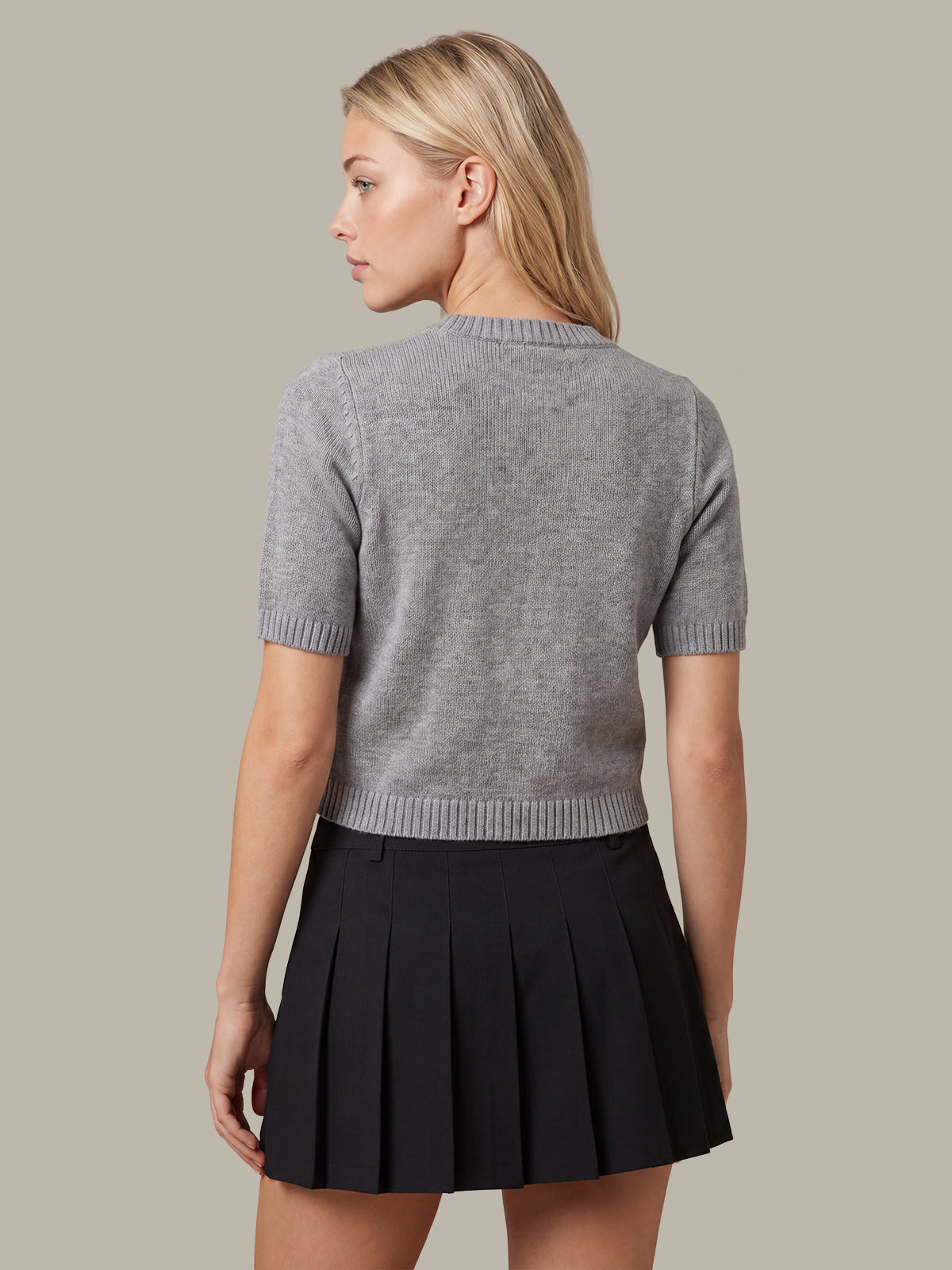 A woman shown from the back wearing a short-sleeve gray knit top and a black pleated mini skirt, against a plain warm gray studio background.