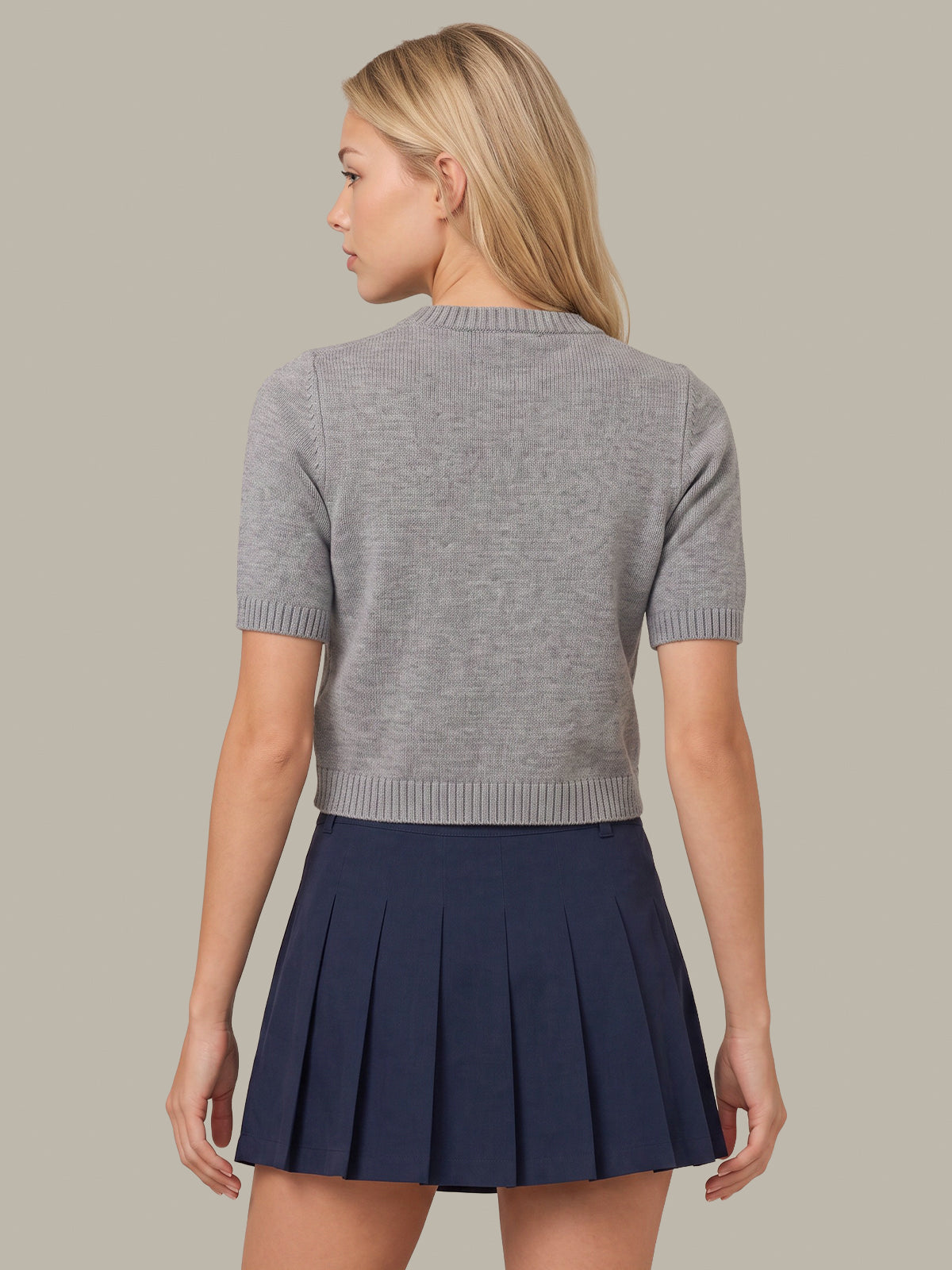 A woman shown from the back wearing a light gray short-sleeve knit top and a navy blue pleated mini skirt, set against a solid warm gray studio background.