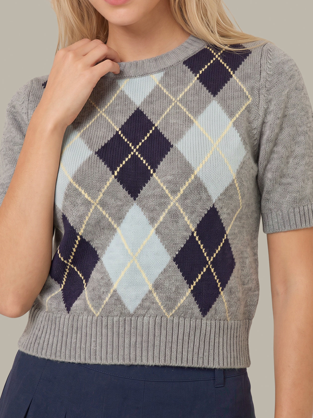 A close-up of a woman wearing a gray short-sleeve knit sweater with an argyle pattern in navy, light blue, and cream, paired with navy bottoms.