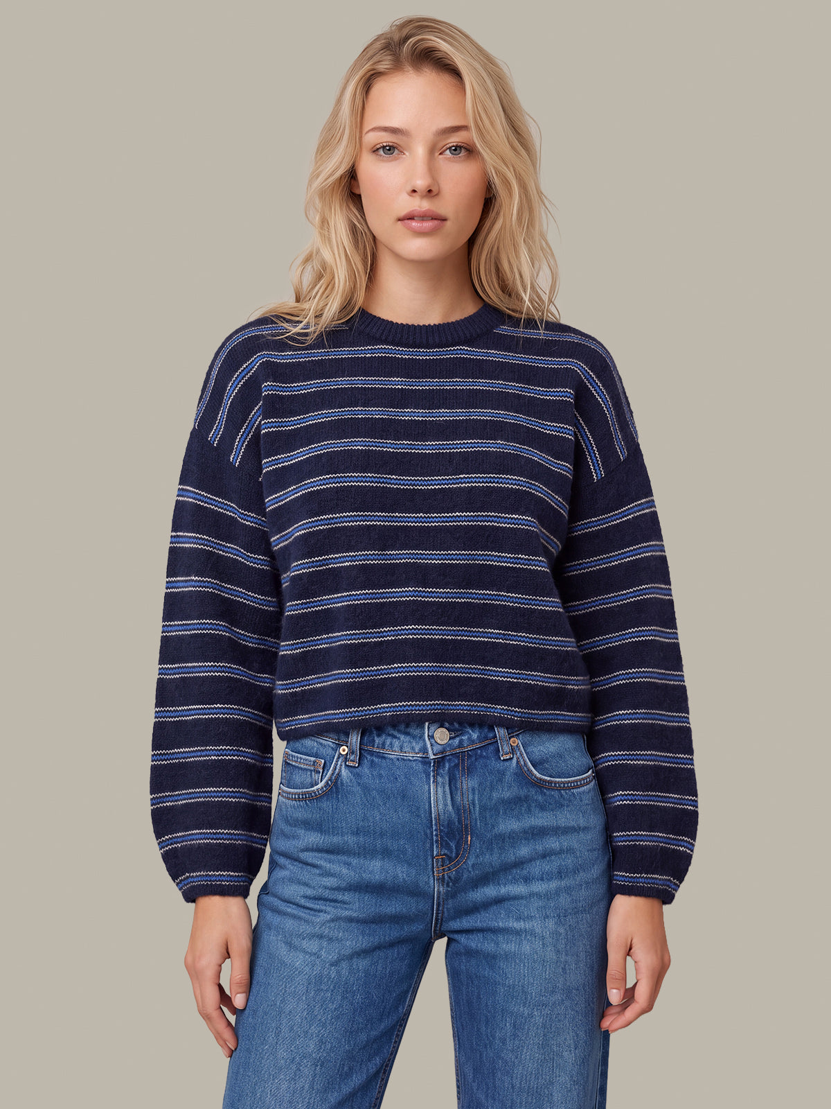A woman modeling a navy blue knit sweater with thin horizontal light blue and white stripes, paired with blue jeans, against a solid taupe background.