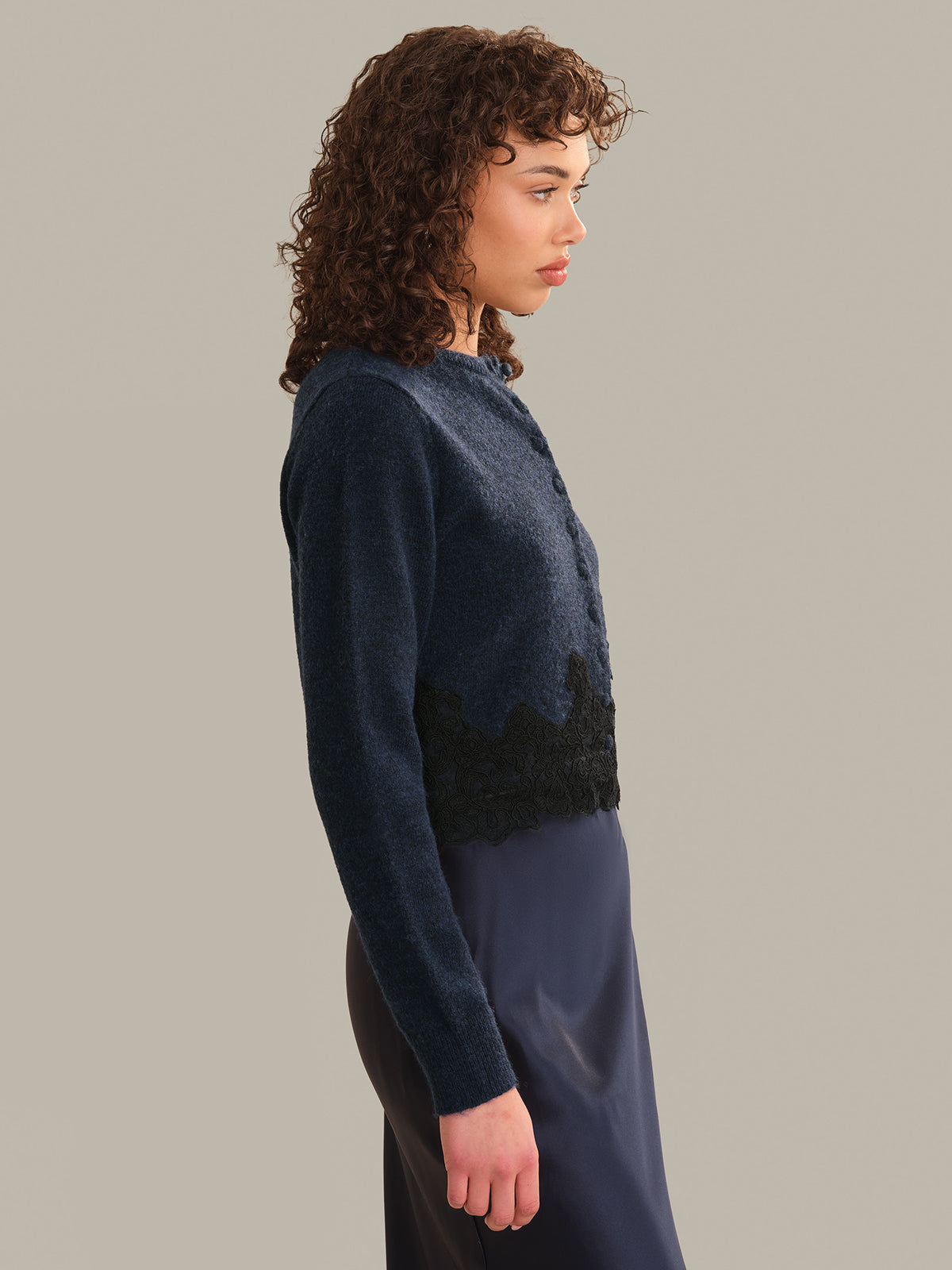 A model shown in profile wearing a dark navy blue knit cardigan with a black textured lace-like hem detail, paired with a matching dark skirt, against a solid light gray studio background.