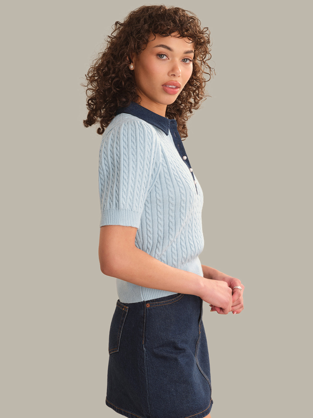 Woman wearing a light blue knitted polo shirt and dark denim skirt on a plain background