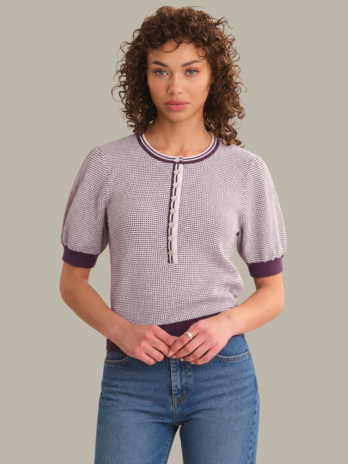 A woman with curly brown hair wearing a short-sleeve knit top with a fine black and white micro-pattern, dark purple trim, and a front button placket, paired with blue jeans, against a solid light gray background.