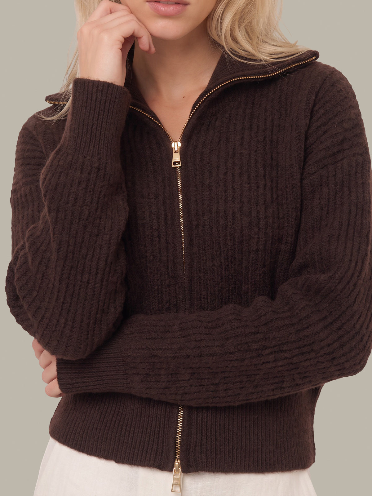 A woman wearing a dark brown ribbed knit zip-up sweater with a high collar and gold-tone zipper, shown from the shoulders to the hips against a plain light gray background.
