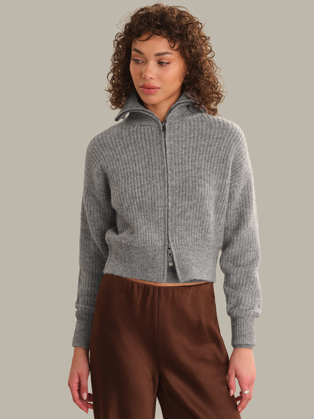 A model with curly brown hair wearing a gray ribbed knit zip-up hoodie and brown pants, photographed against a solid light gray background.
