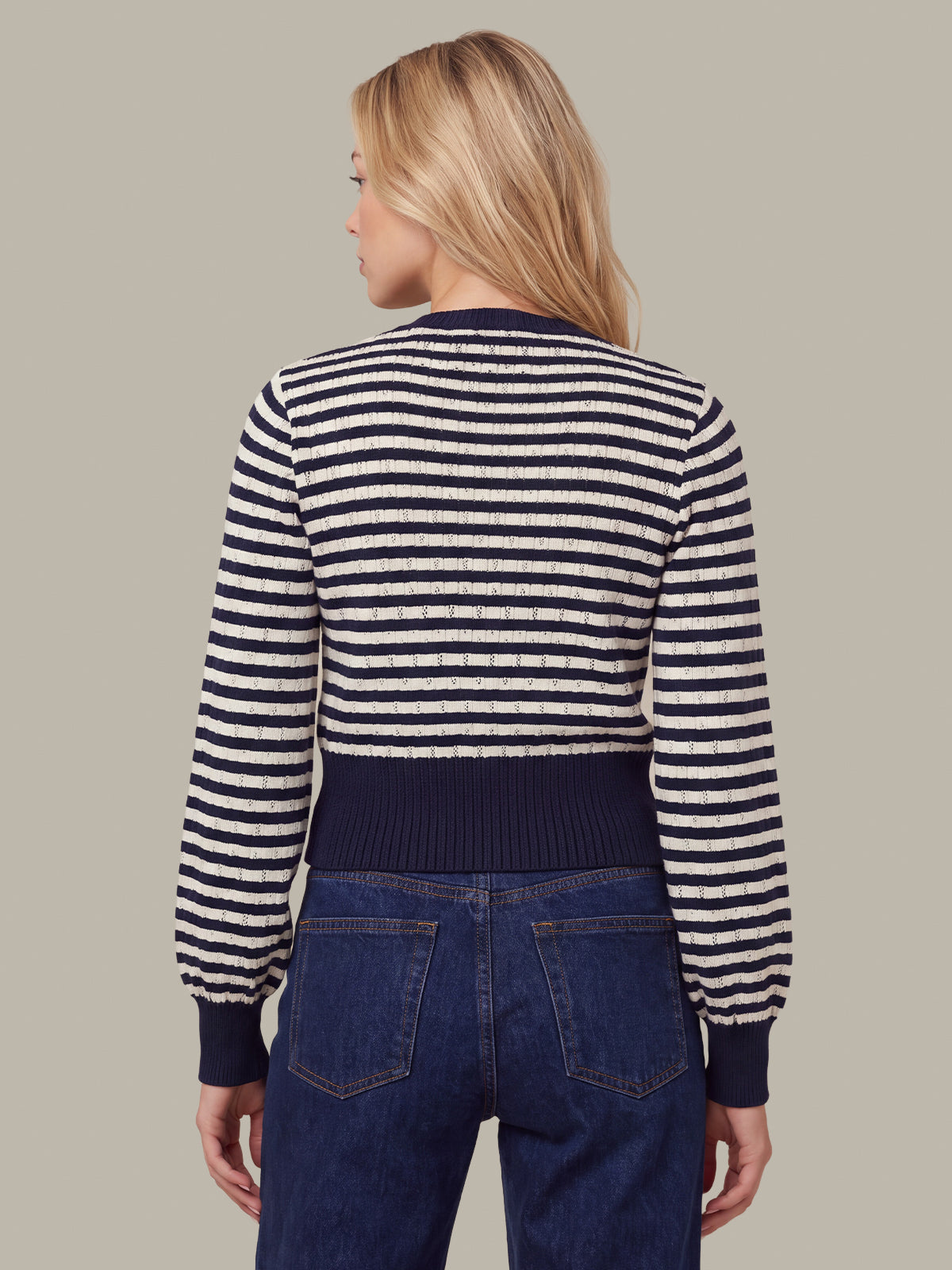 A woman shown from the back wearing a navy and cream horizontal striped knit sweater with dark navy ribbed trim, paired with dark blue jeans, against a solid beige background.